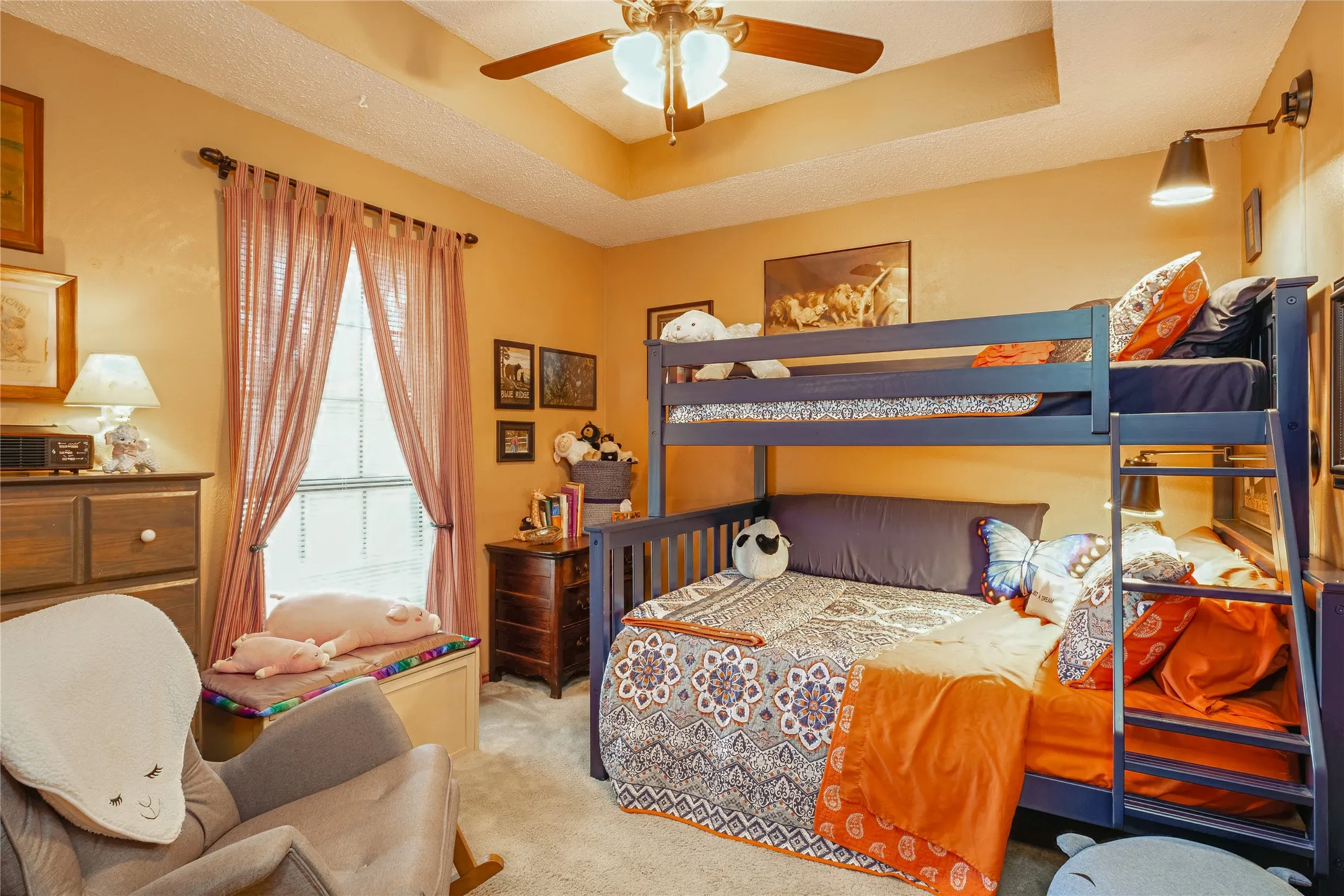 Tray ceiling with a ceiling fan, wall-mounted light fixtures, and a window with blinds