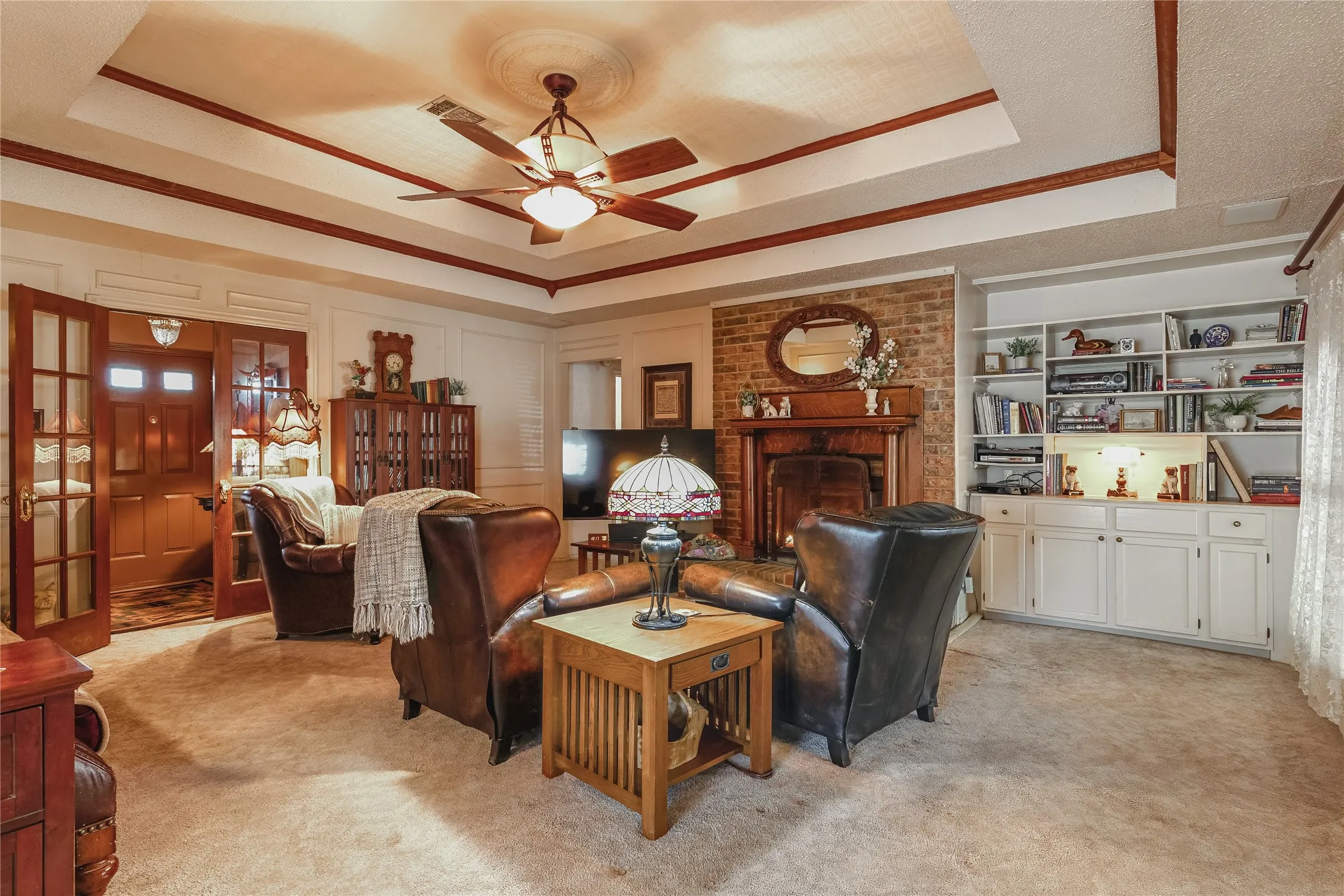 Recessed ceiling with wood-finish trim, ceiling fan with integrated lighting, brick-clad fireplace with wood-finish mantel, built-in shelving, and built-in cabinetry