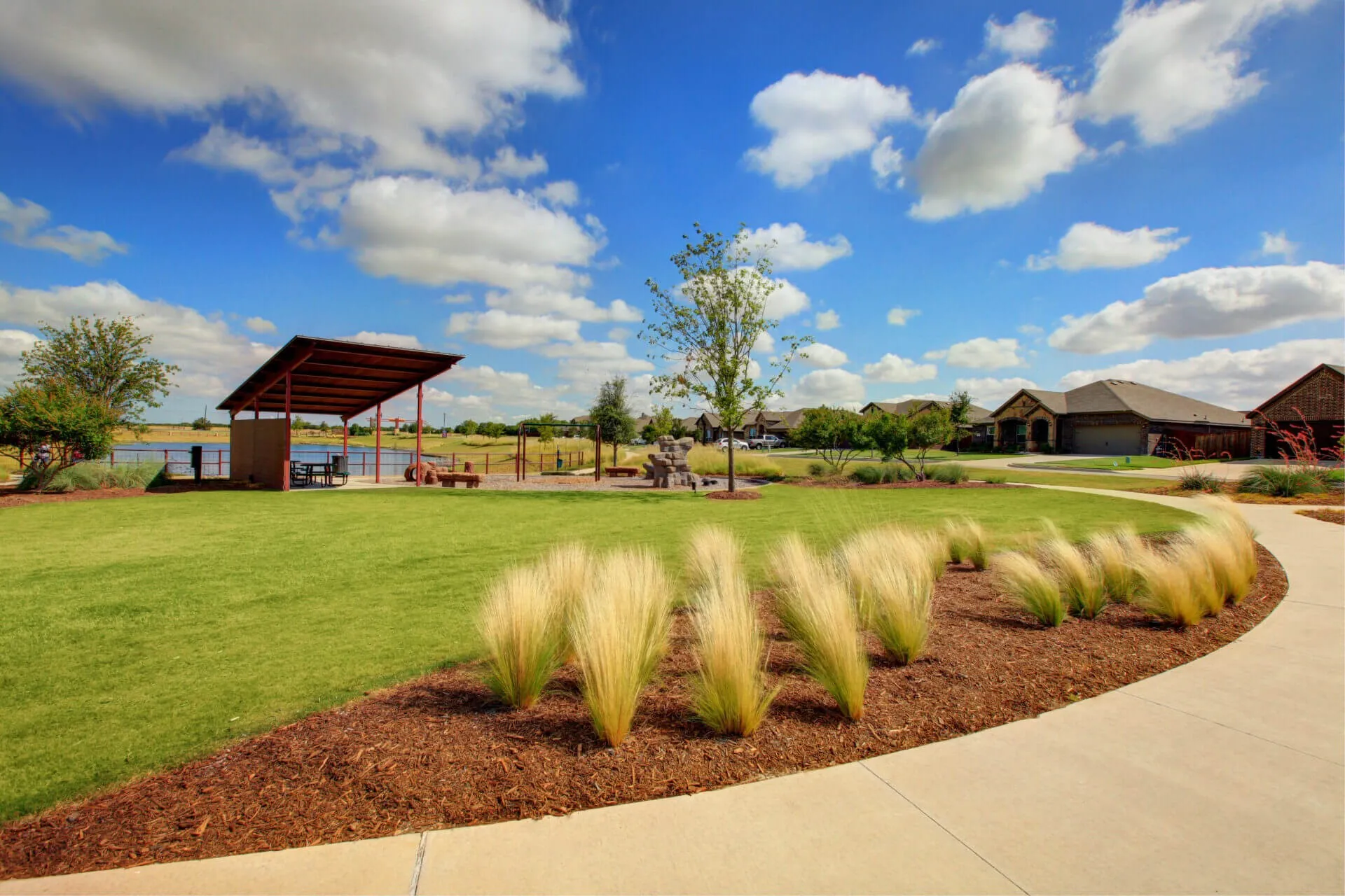Manicured community green space featuring a paved pathway, decorative landscaping, and a covered pavilion with lake access