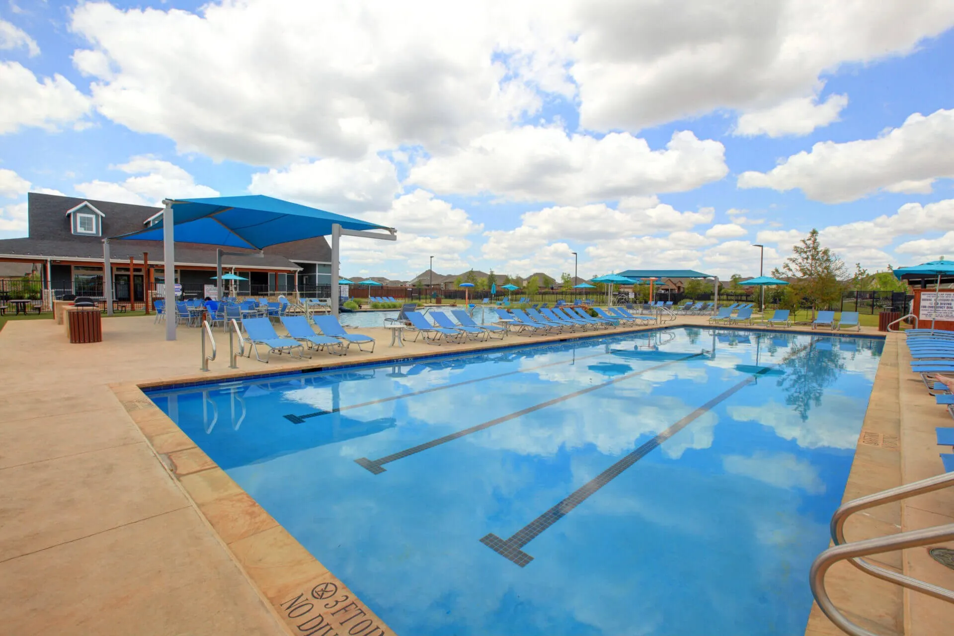 Resort-style pool area featuring lap lanes, a spacious concrete deck, shaded seating, numerous lounge chairs, and an adjacent clubhouse