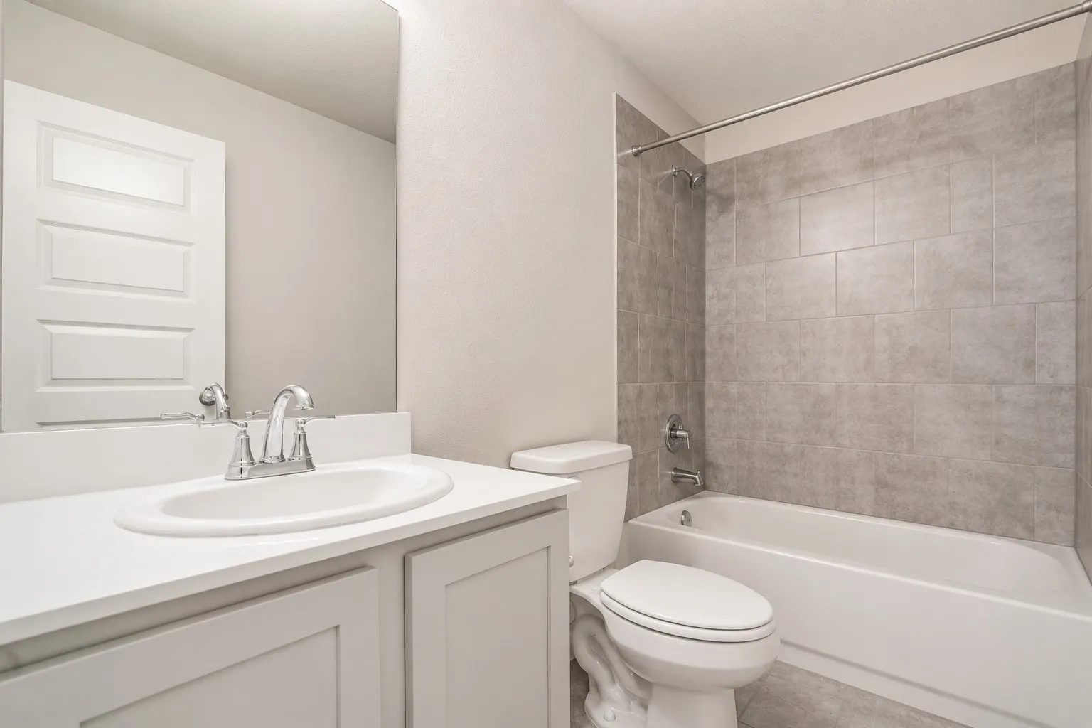 White vanity with an integrated sink, polished chrome faucet, full-width mirror, toilet, and bathtub with gray tile surround