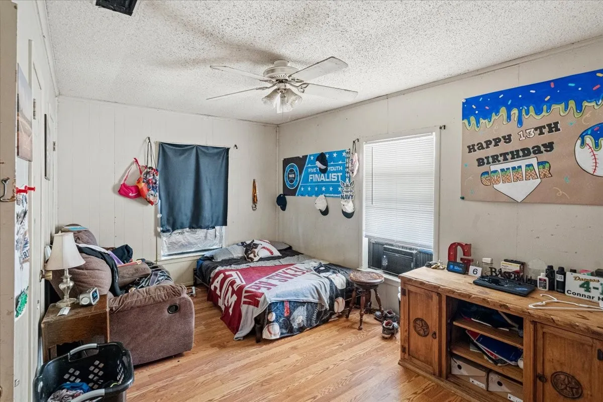 Wood-finish flooring throughout, featuring a ceiling fan, two windows with blinds, and paneled walls