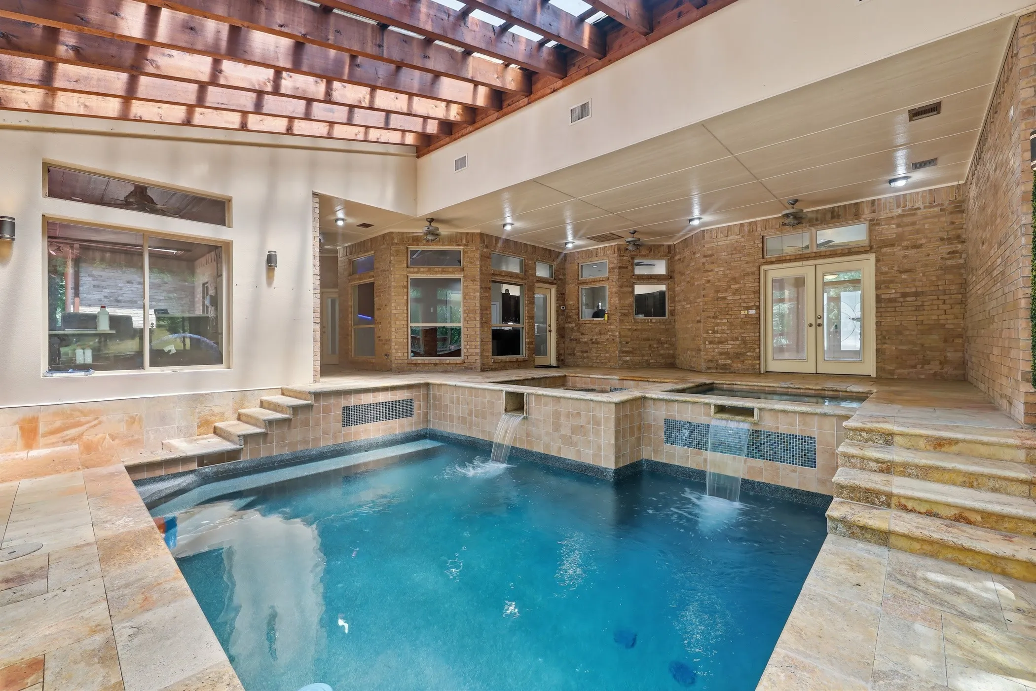 Indoor pool with integrated spa and waterfall features, surrounded by stone tile decking