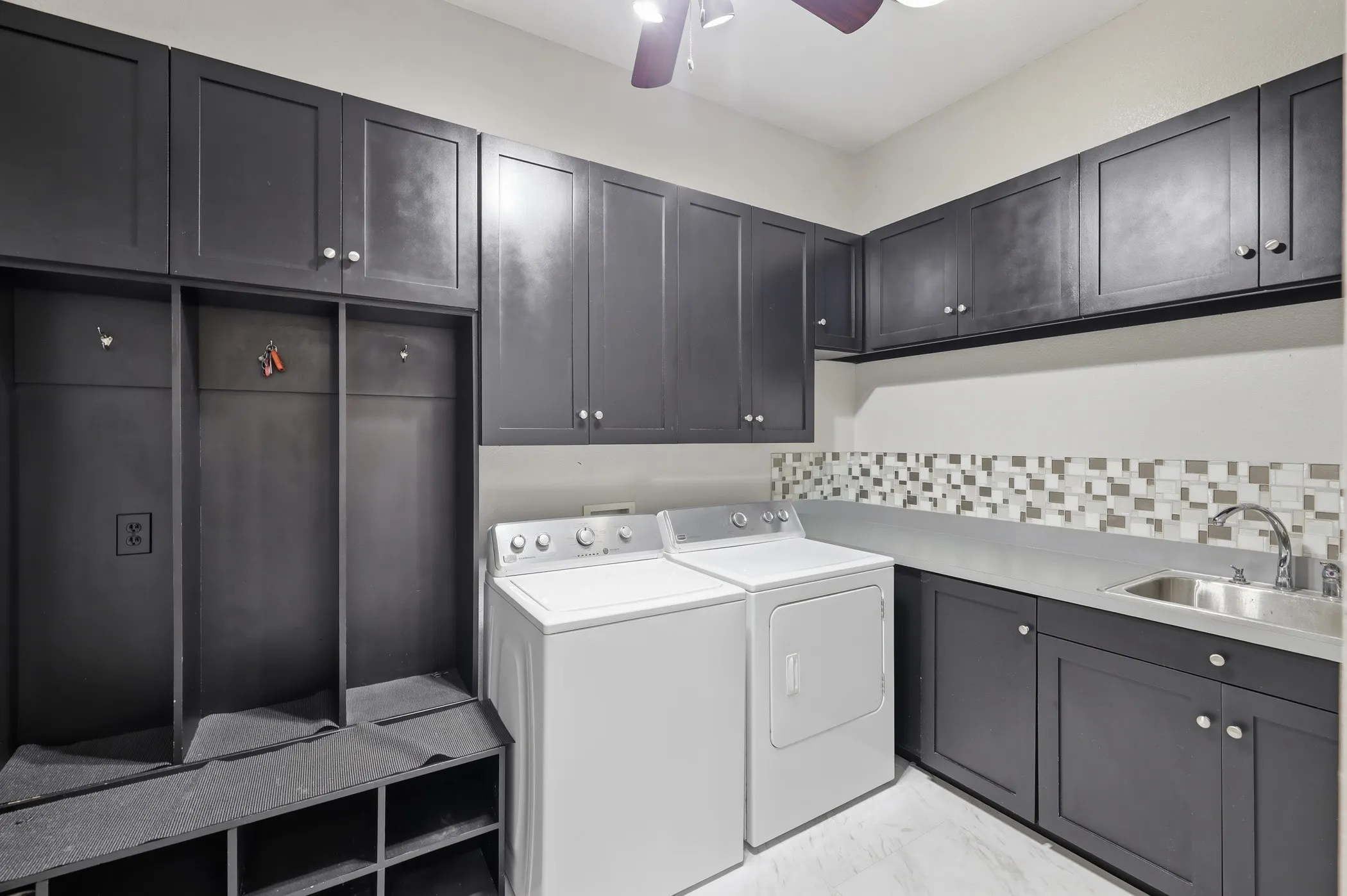 Laundry room featuring dark cabinetry, built-in storage, a utility sink, mosaic tile backsplash, and light-toned flooring