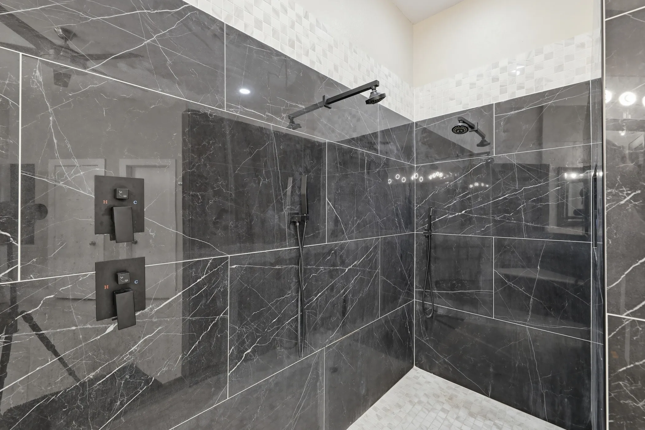 Spacious shower featuring large format dark tile with white veining, dual matte black shower heads, a handheld shower fixture, and a mosaic tile floor