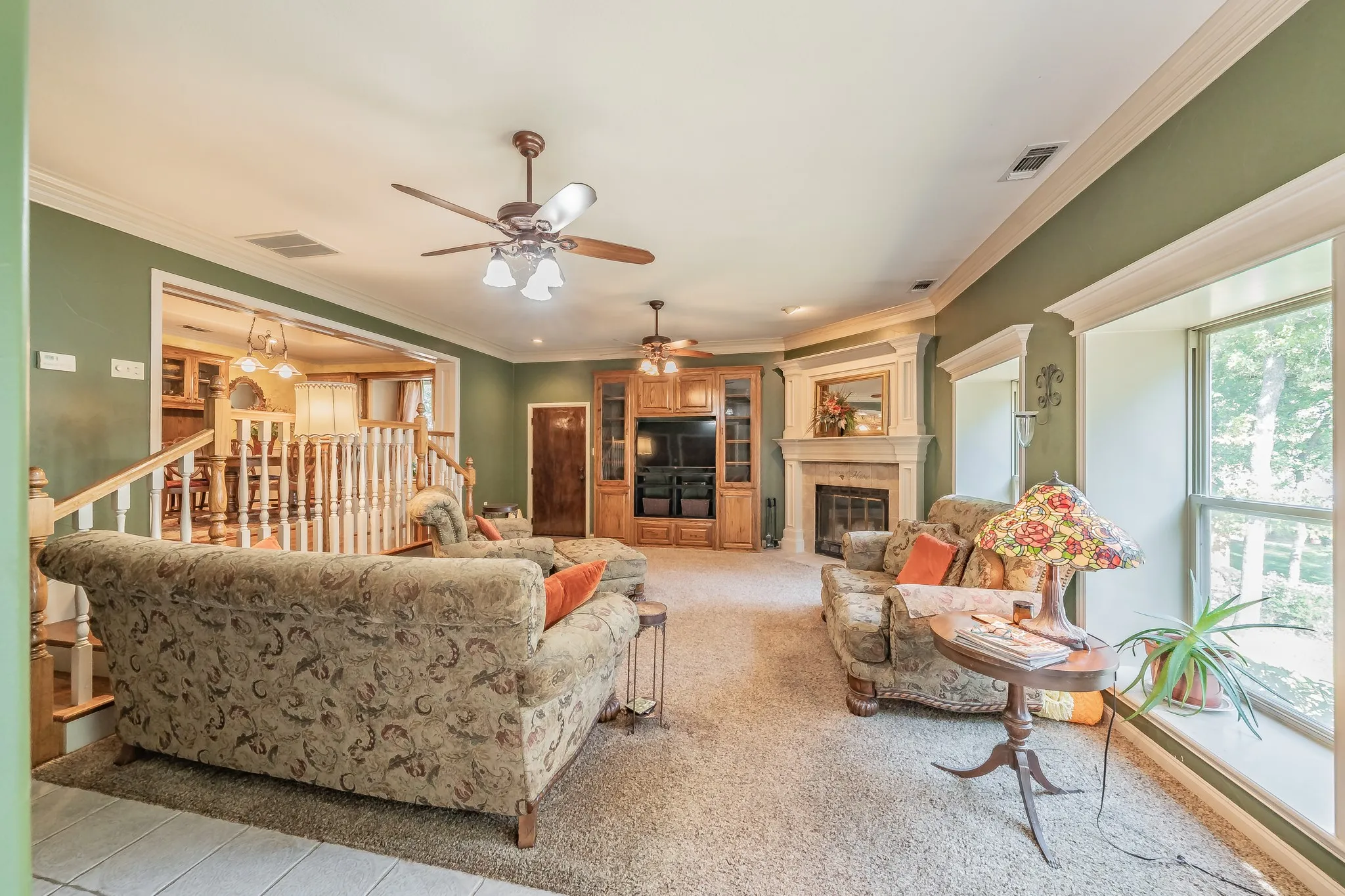 Living room with a glass covered wood buringfireplace, ornamental molding, ceiling fan, light colored carpet, and light tile patterned floors