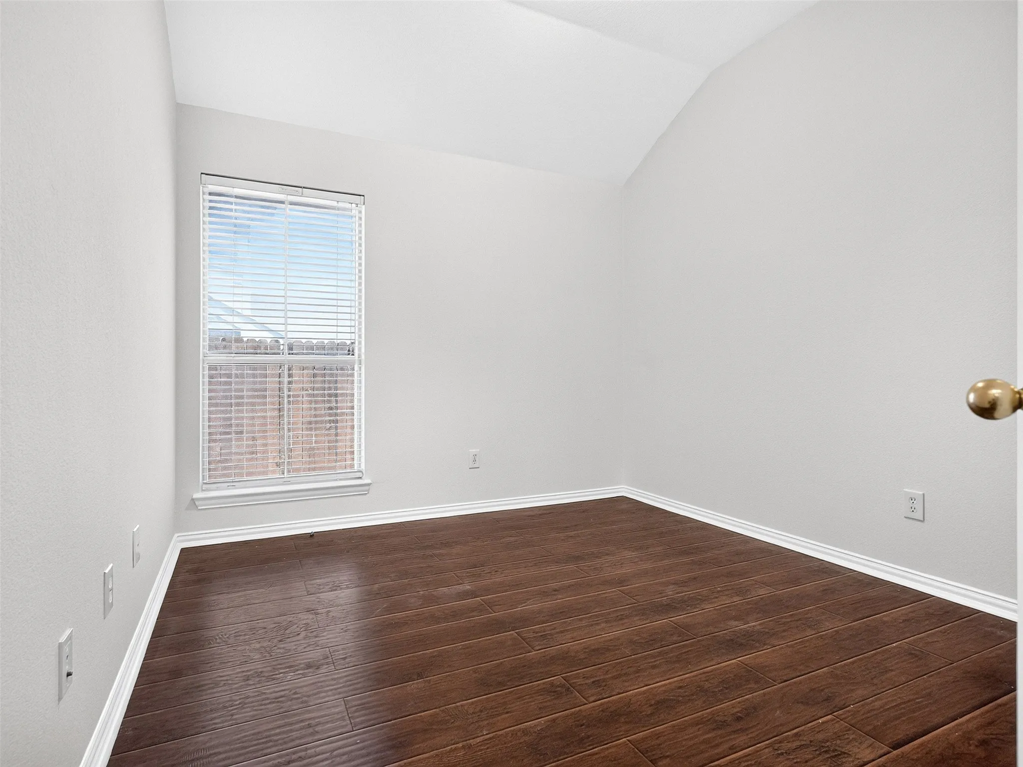 Vaulted ceiling, wood-finish flooring, single window with blinds, white trim, and a brass door handle
