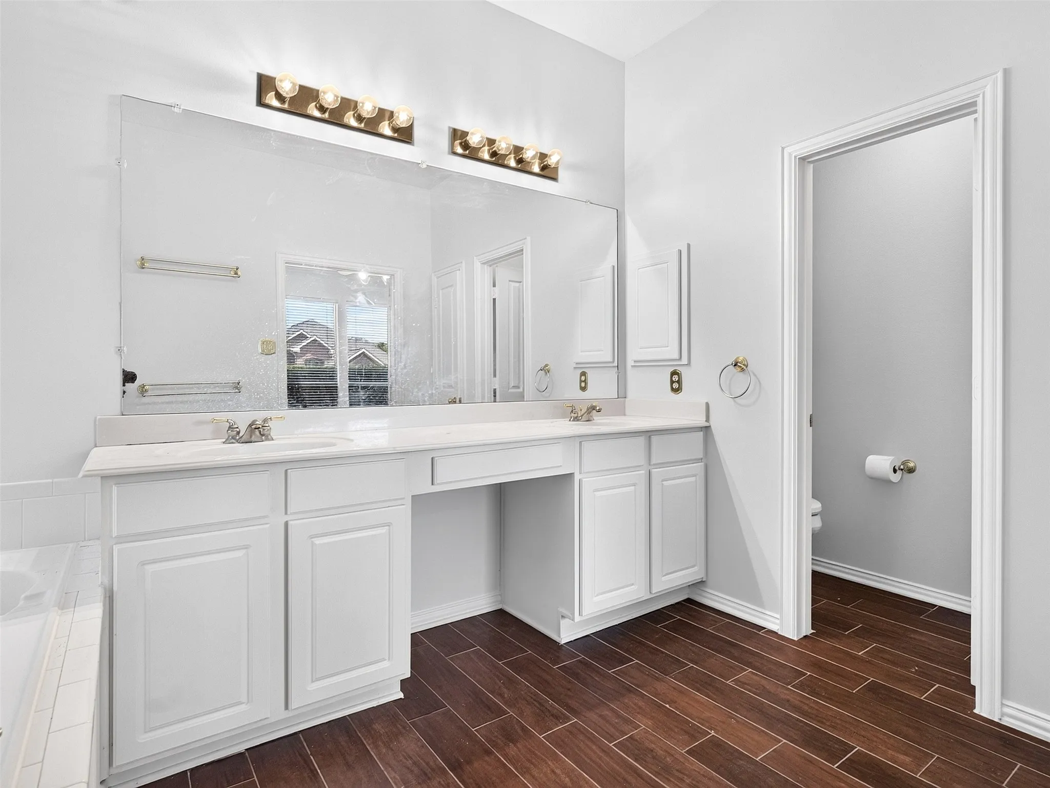 Dual white vanities with integrated sinks, expansive wall-mounted mirror, vanity lighting fixtures, wood-finish flooring, and private water closet