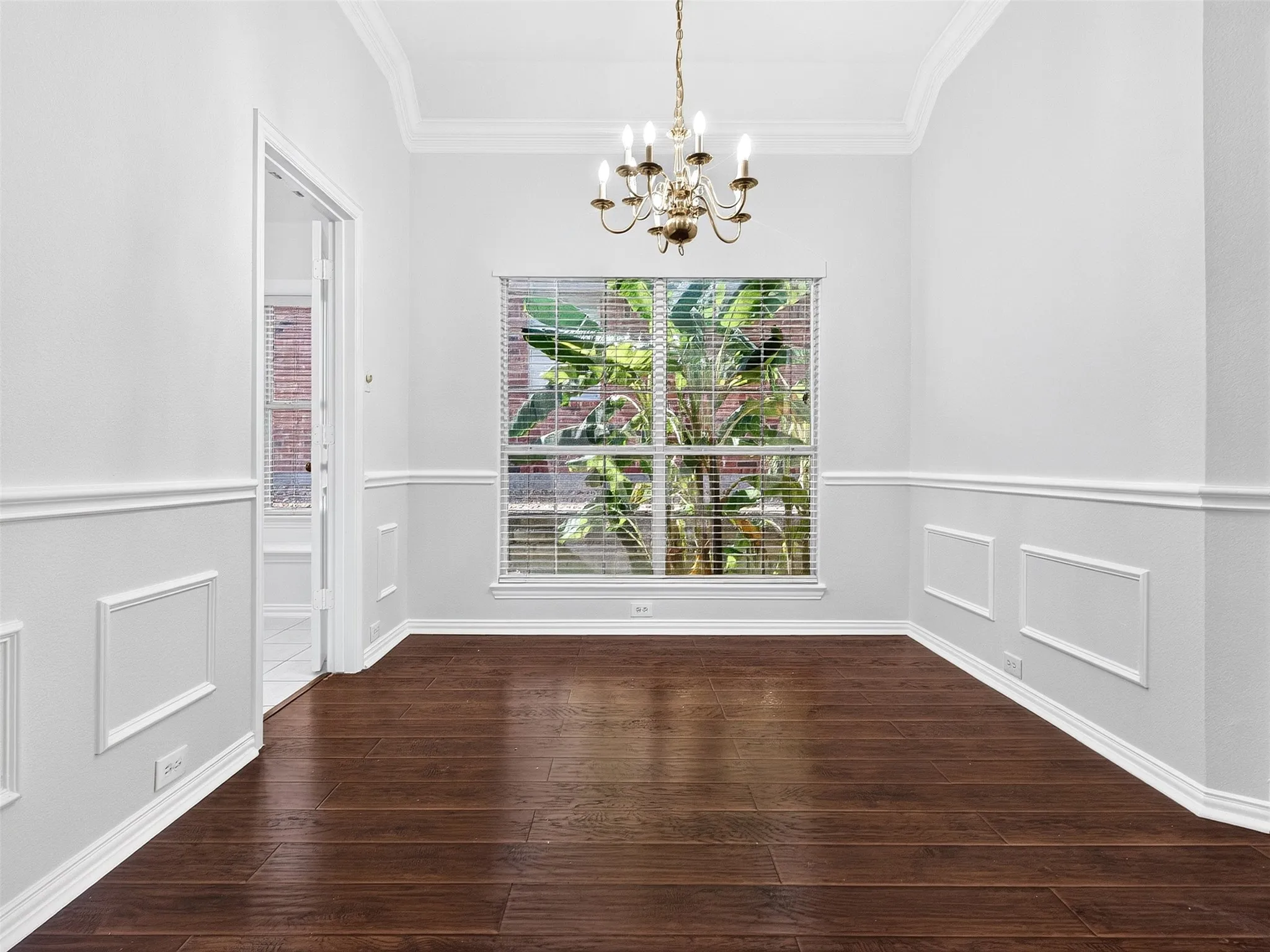Wood-finish flooring, wainscoting, crown molding, a window with blinds, and a brass chandelier