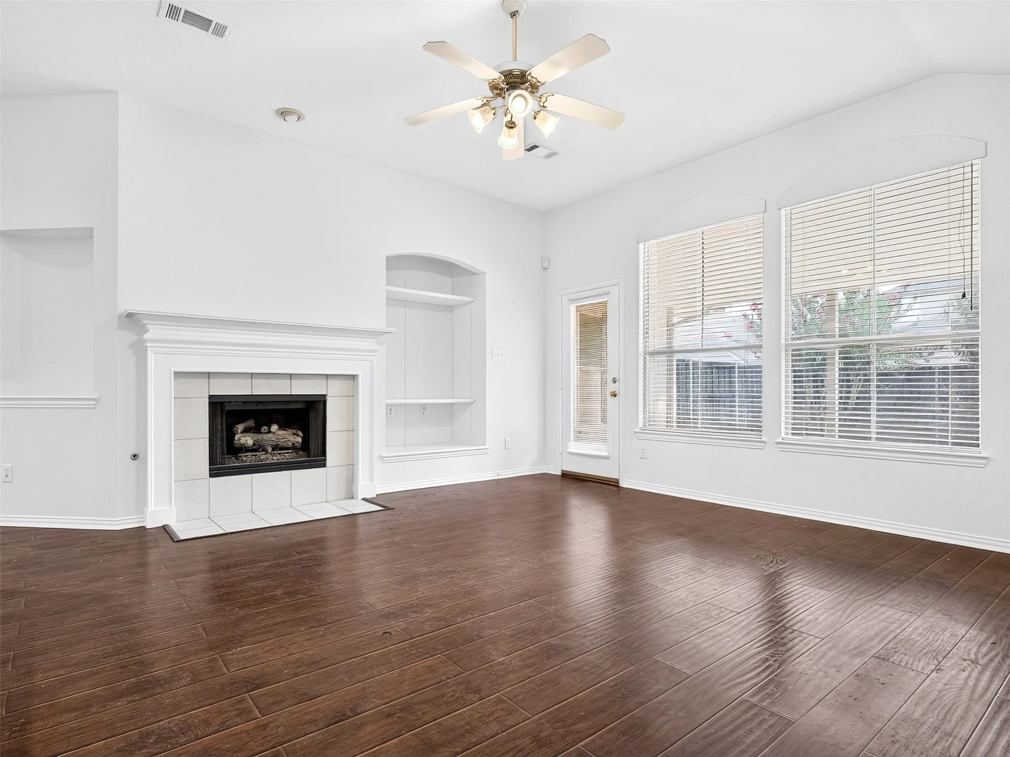 Wood-finish flooring, white tile fireplace with mantle, built-in shelving, two large windows with blinds, and ceiling fan with lighting