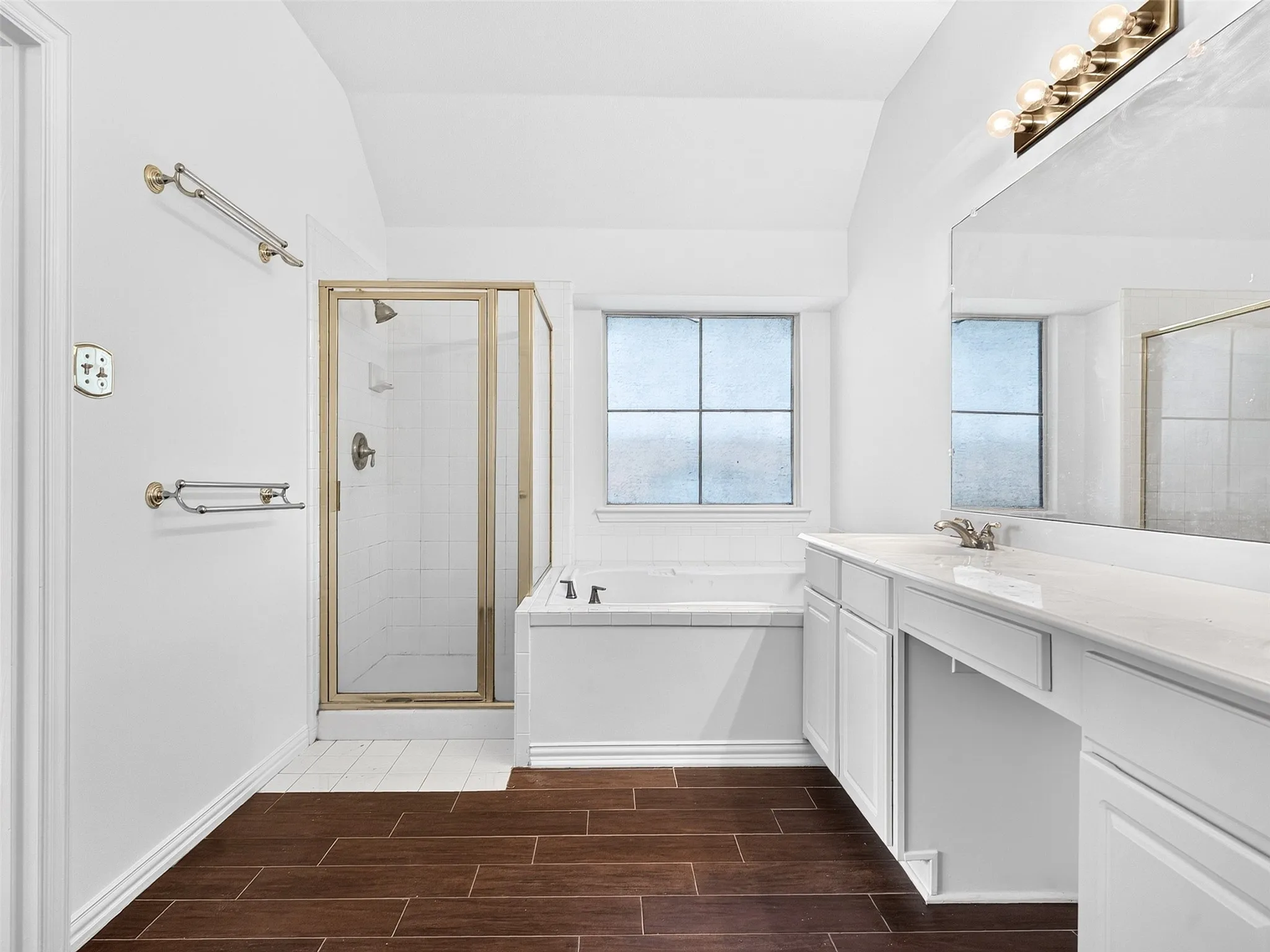Vaulted ceiling, wood-finish flooring, white vanity with integrated sink, walk-in shower with glass enclosure, and separate soaking tub