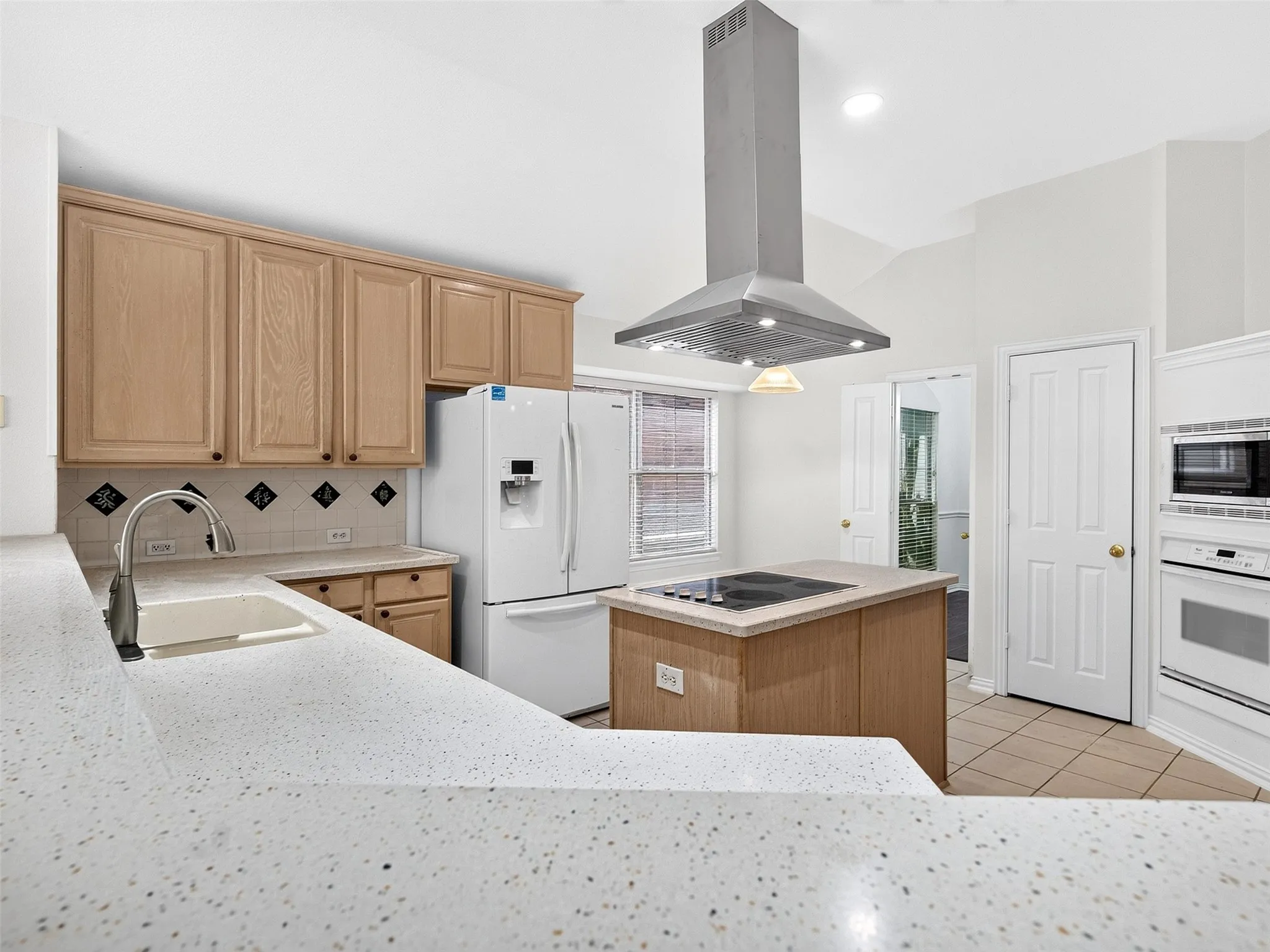 Kitchen with light wood-finish cabinetry, white solid surface countertops, stainless steel range hood, and a built-in oven and microwave