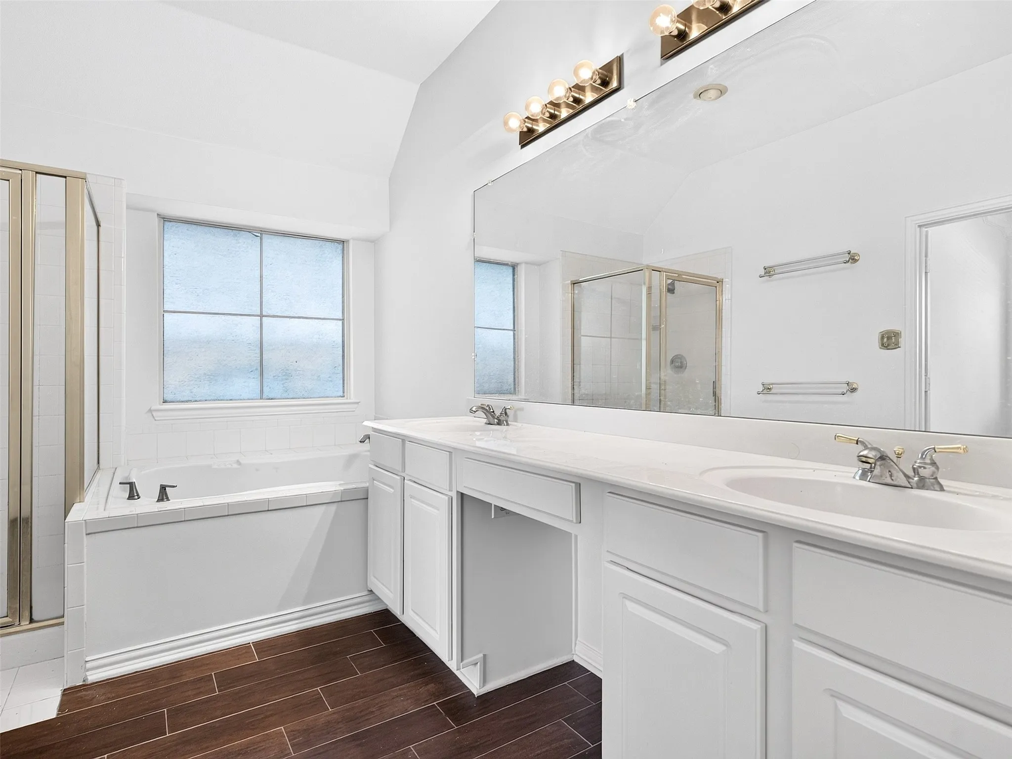 Dual vanity with white cabinetry and integrated sinks, a built-in tub with tile surround, a glass-enclosed shower, and wood-finish flooring