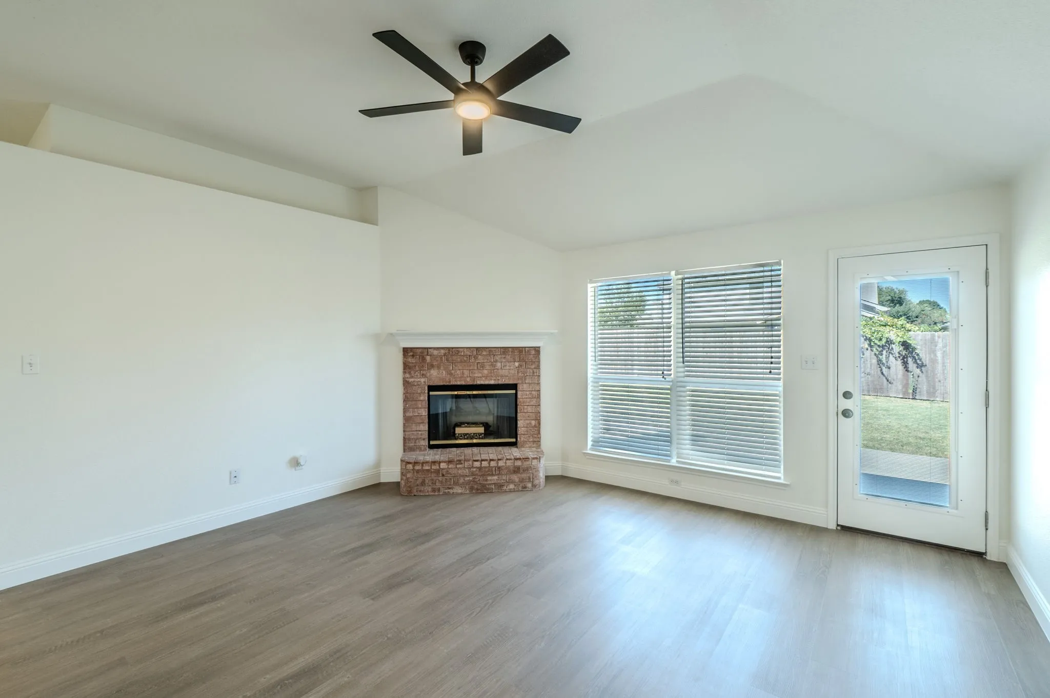 Vaulted ceiling with a ceiling fan, wood-finish flooring, brick-surround fireplace, and a glass-paneled exterior door