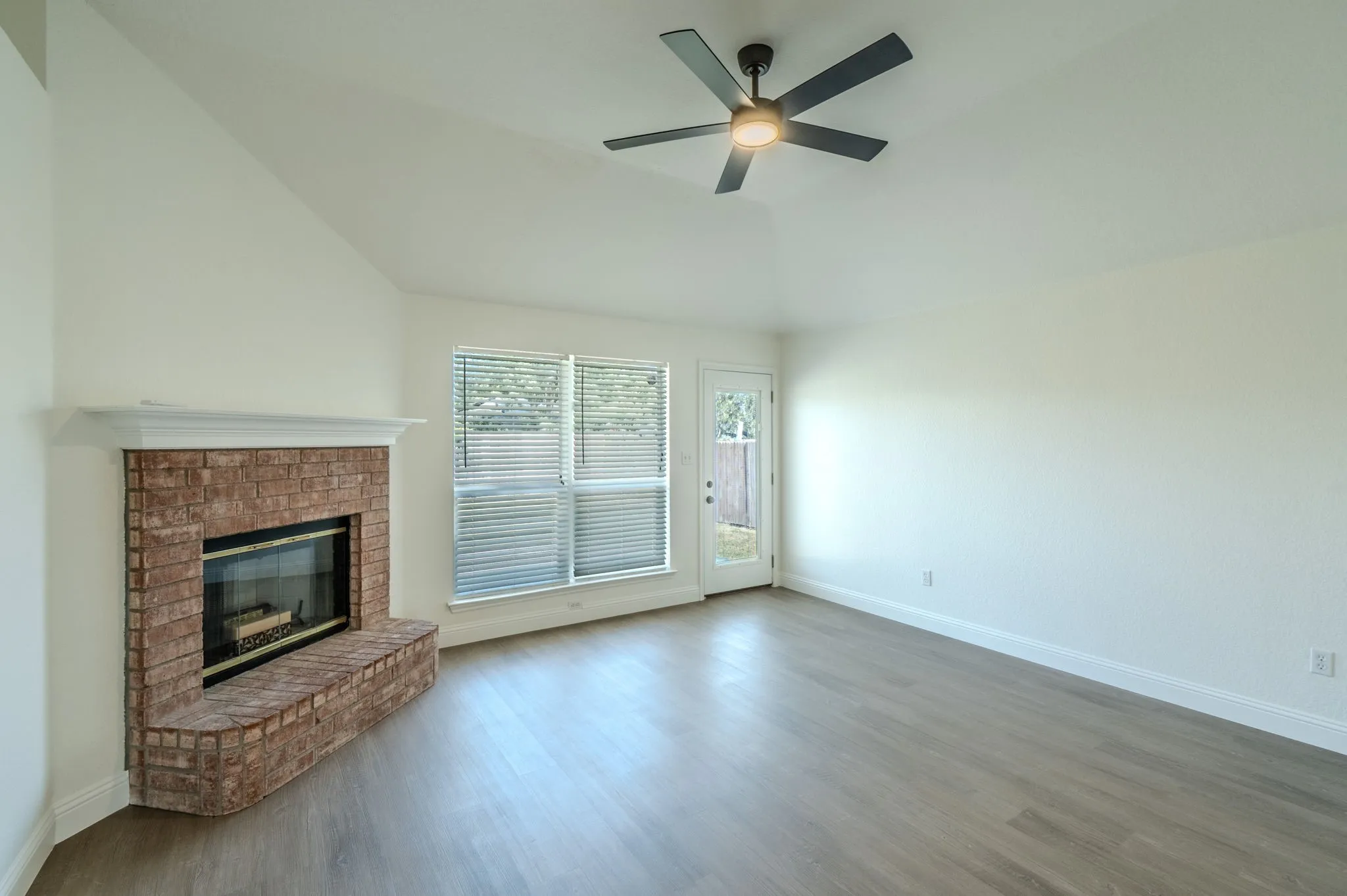 Vaulted ceiling, contemporary ceiling fan, brick fireplace with mantel, wood-finish flooring, and exterior access via glass-paned door