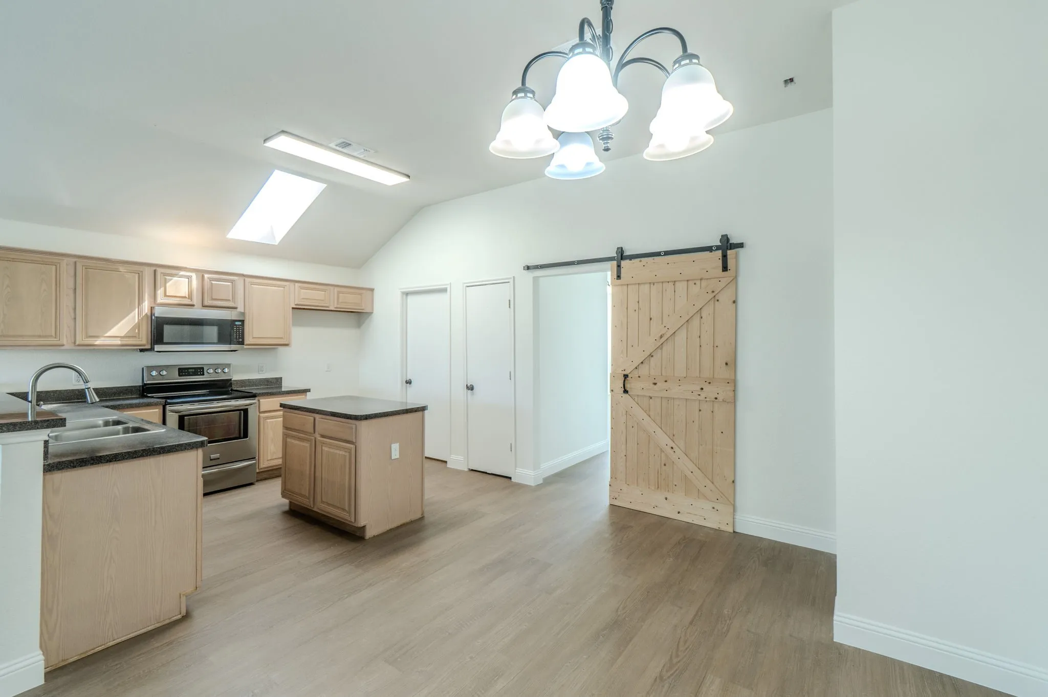Vaulted ceiling with skylight, wood-finish flooring, stainless steel appliances, kitchen island, and barn door