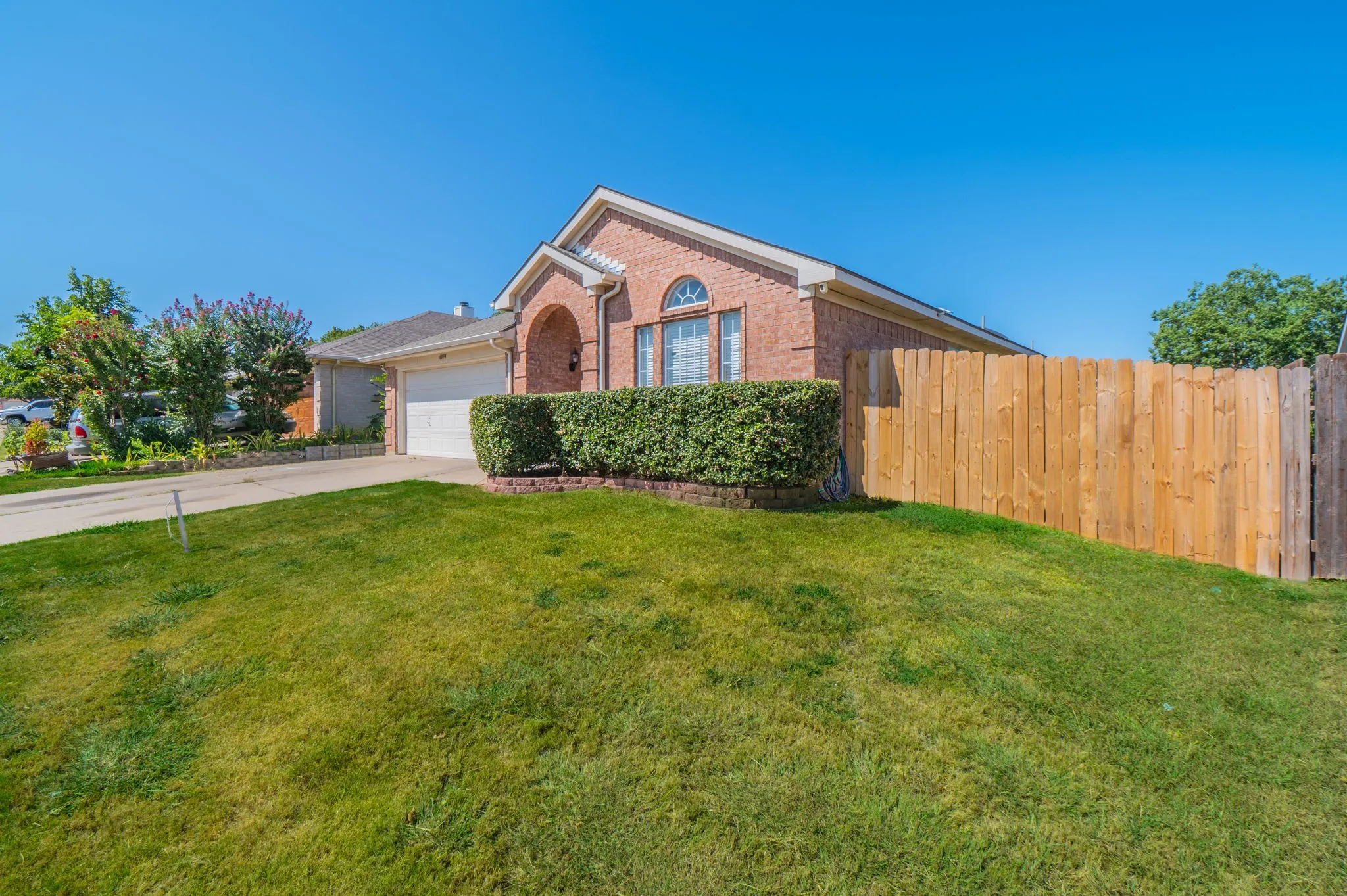 Brick exterior, arched entryway, attached garage, manicured shrubbery, and a wooden privacy fence