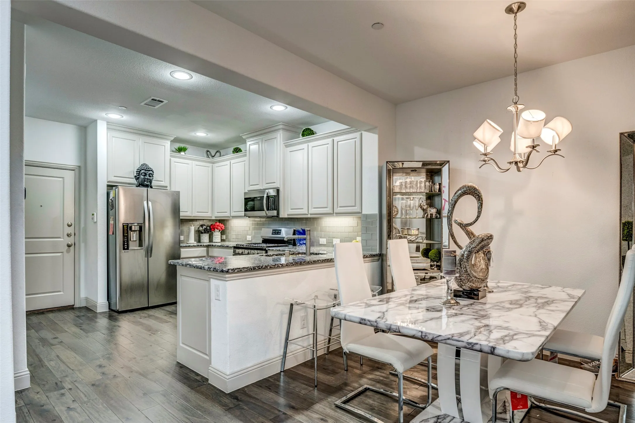 Open-concept kitchen and dining area featuring wood-finish flooring, white cabinetry, stainless steel appliances, granite countertops, and a marble-finish dining table