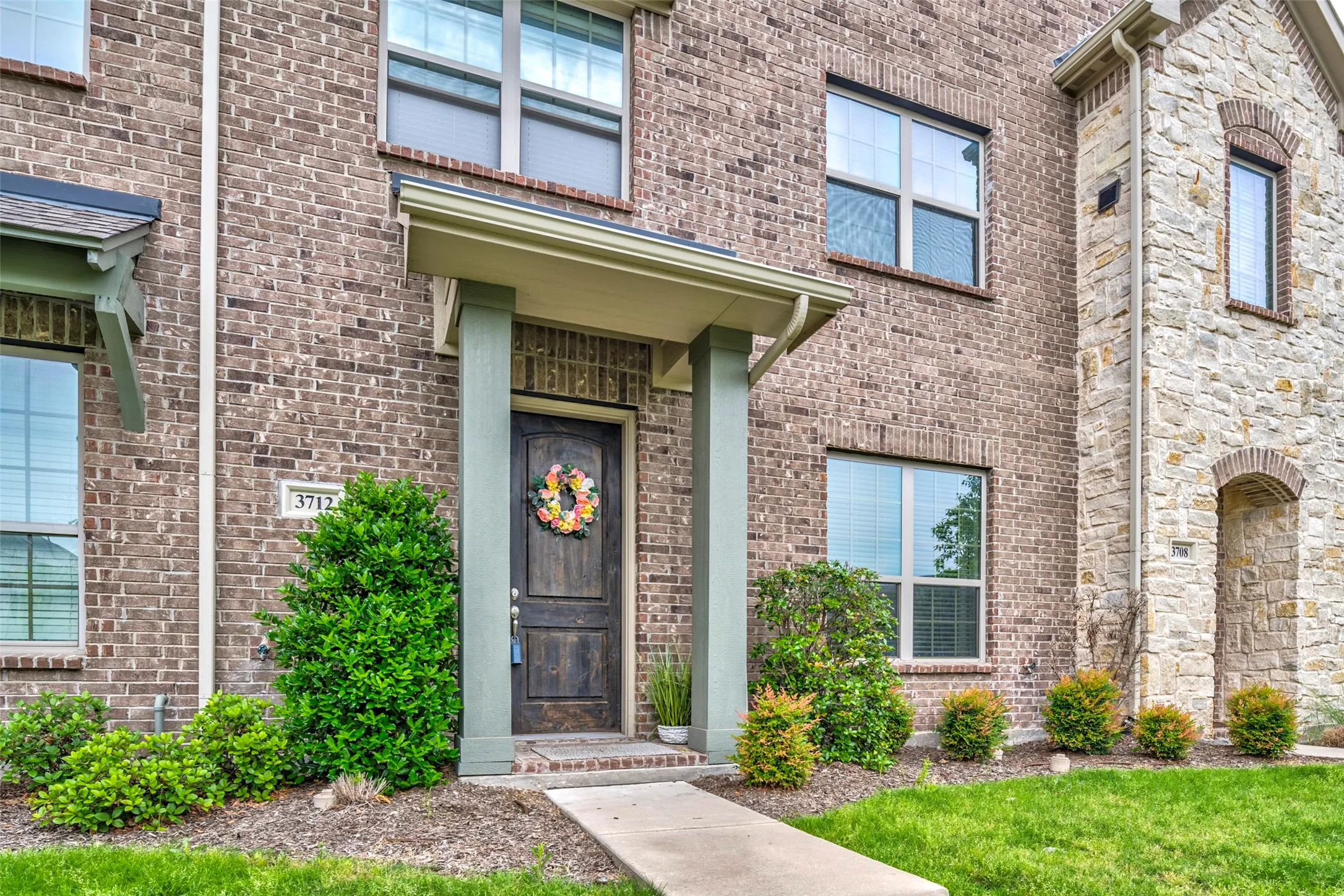 Brick and stone facade featuring a wood entry door with an overhead portico, multiple windows with blinds, a concrete walkway, and landscaped grounds with mature shrubbery and a manicured lawn