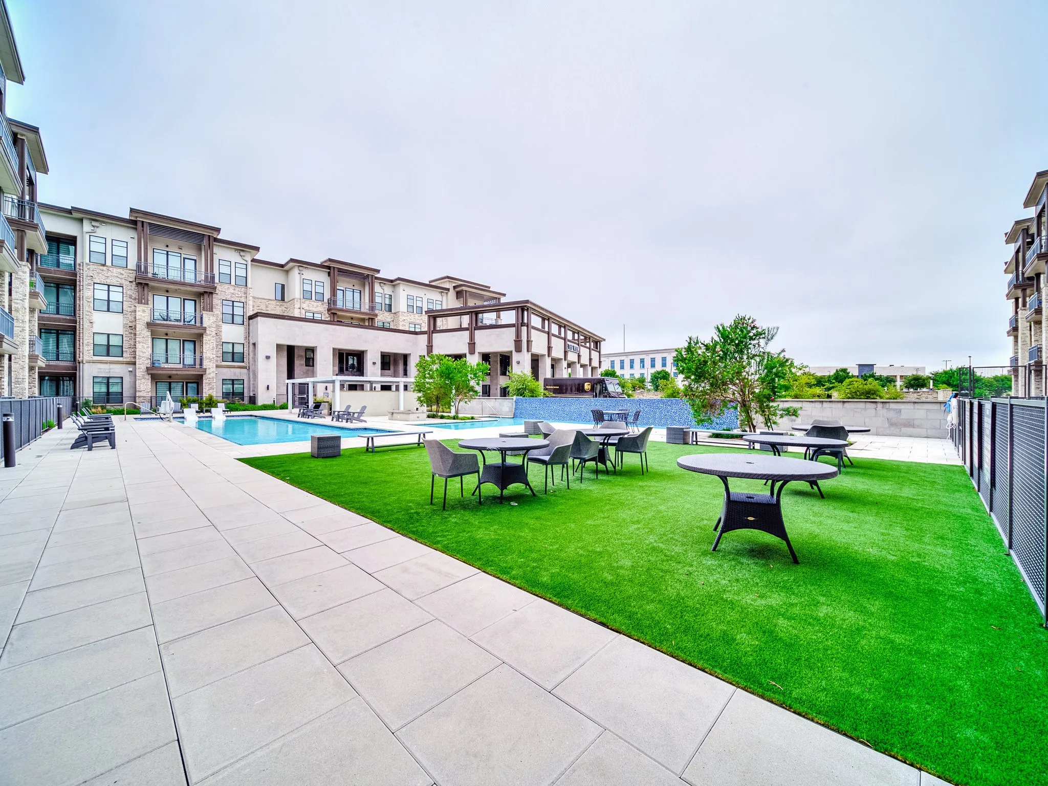 Expansive outdoor space featuring a rectangular swimming pool, paver stone hardscaping, artificial turf lawn, and a multi-story building exterior with stone and siding accents