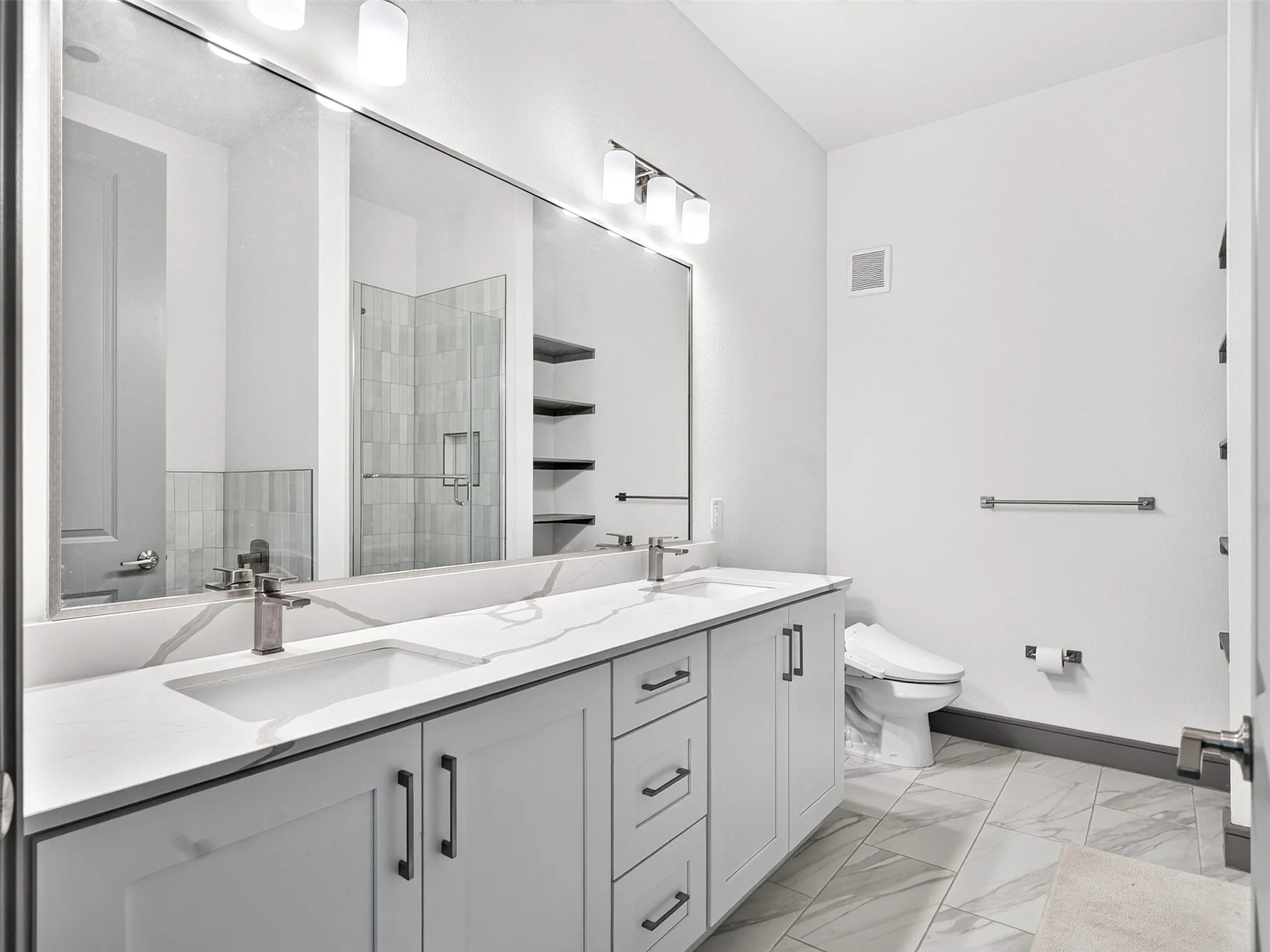 Dual vanity with integrated sinks, expansive mirror, light-toned tile flooring, contemporary light fixtures, and a shower enclosure visible in reflection