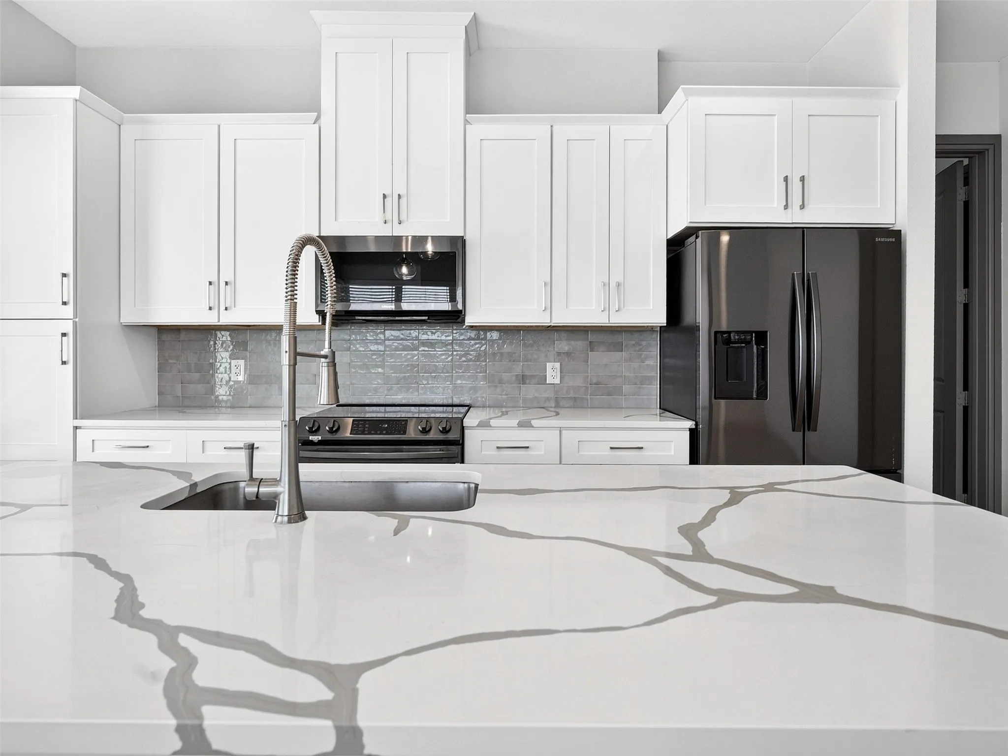 Kitchen featuring white shaker-style cabinetry, a large kitchen island with a white countertop and gray veining, stainless steel sink with a high-arc faucet, and dark gray appliances