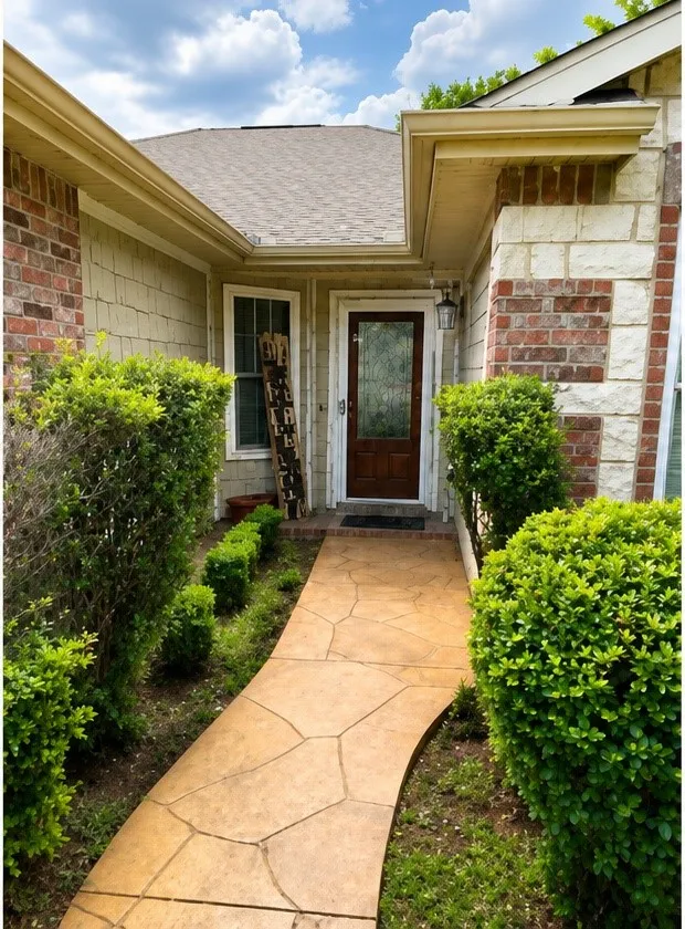 Textured concrete pathway, brick and stone exterior accents, dark wood entry door with decorative glass, and established perimeter landscaping