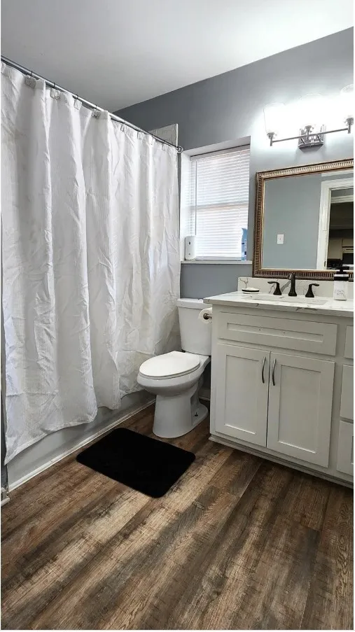 Wood-finish flooring, white vanity with cabinet storage, countertop with integrated sink, black fixtures, and a decorative mirror