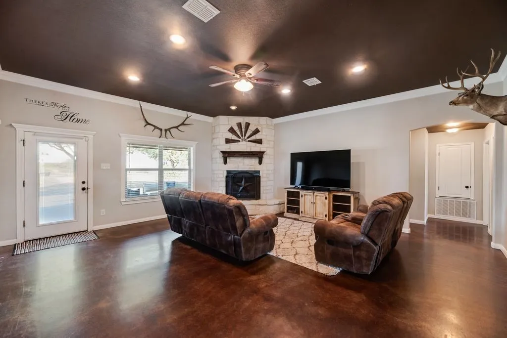 Corner stone fireplace with wood mantel, ceiling fan with integrated lighting, recessed ceiling lighting, and rich concrete flooring