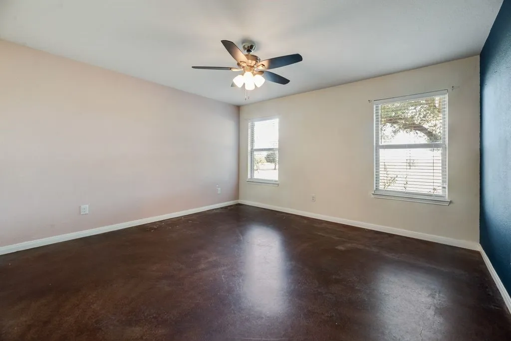 Stained concrete flooring, dual windows with blinds, ceiling fan with integrated lighting, and a contrasting accent wall