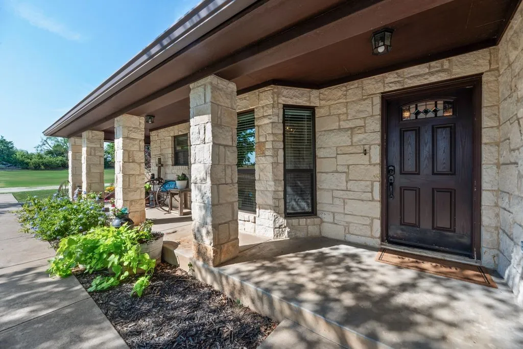 Stone veneer exterior, covered concrete porch, multiple windows with blinds, dark paneled entry door with decorative glass, and exterior lighting fixture