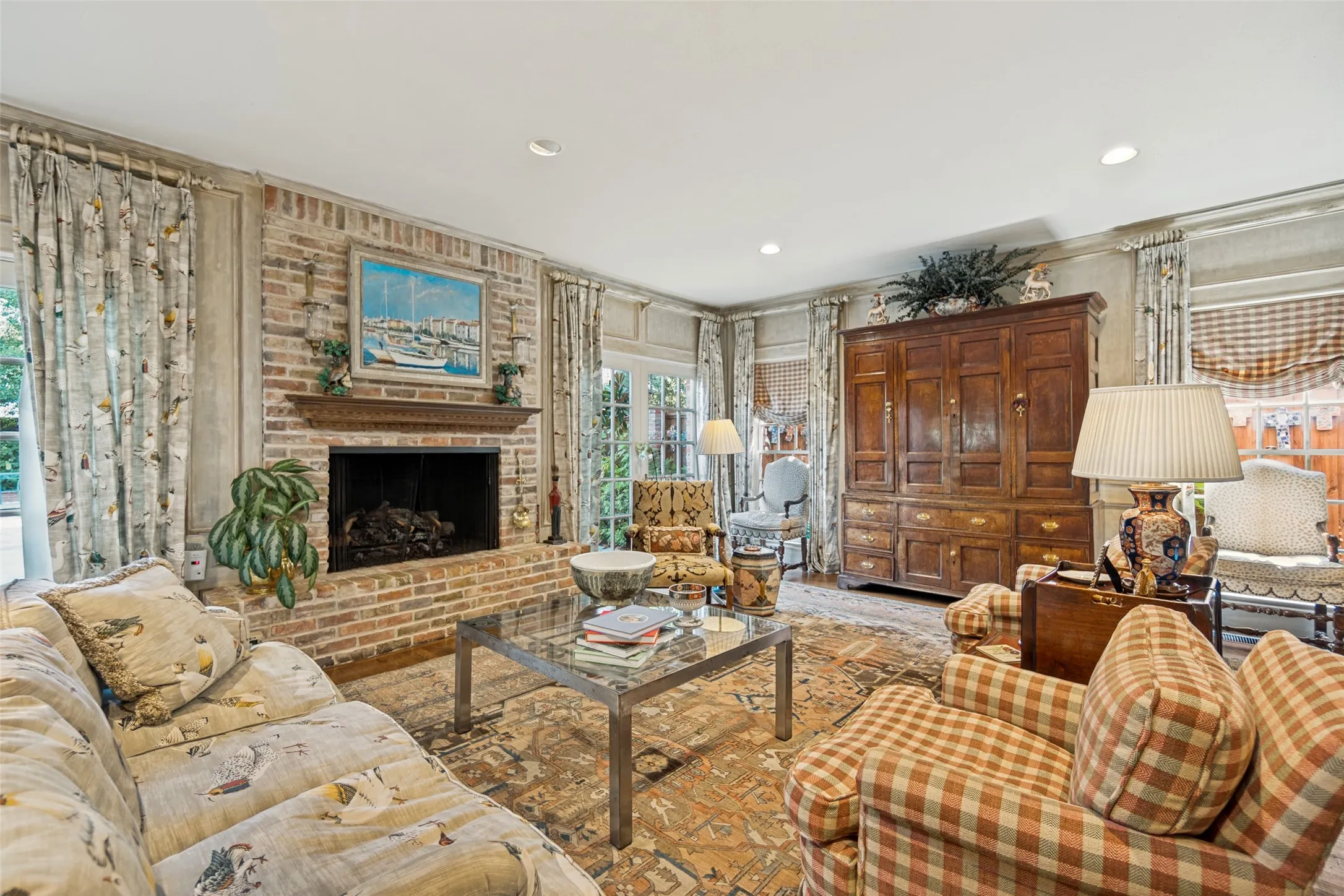 Elegant living room showcasing a brick fireplace with decorative mantel, hardwood flooring, layered drapery with coordinating patterns, recessed ceiling lighting, and substantial wood cabinetry