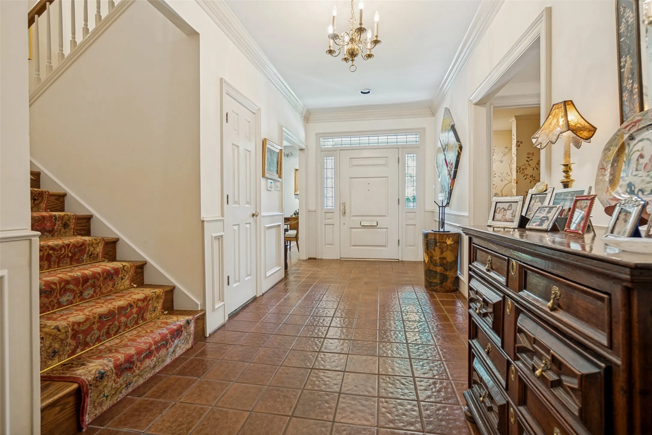 Inviting foyer featuring a chandelier, tile flooring, a paneled front door with sidelights, and a staircase with a decorative runner