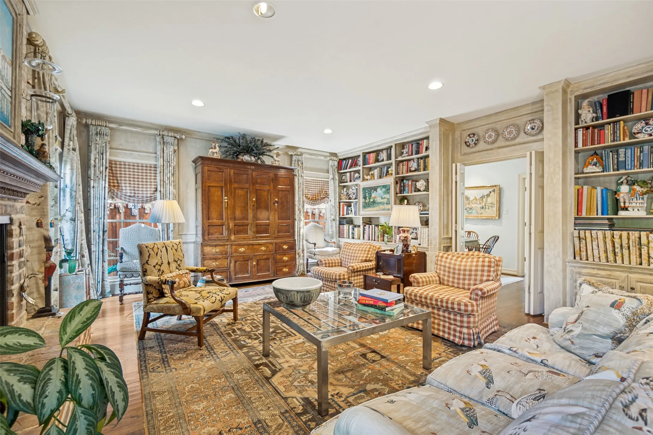 Sophisticated library and sitting room featuring extensive built-in bookshelves with decorative plate display, wood-paneled cabinetry, recessed ceiling lighting, patterned area rug, and multiple seating arrangements anchored by a glass-top coffee table