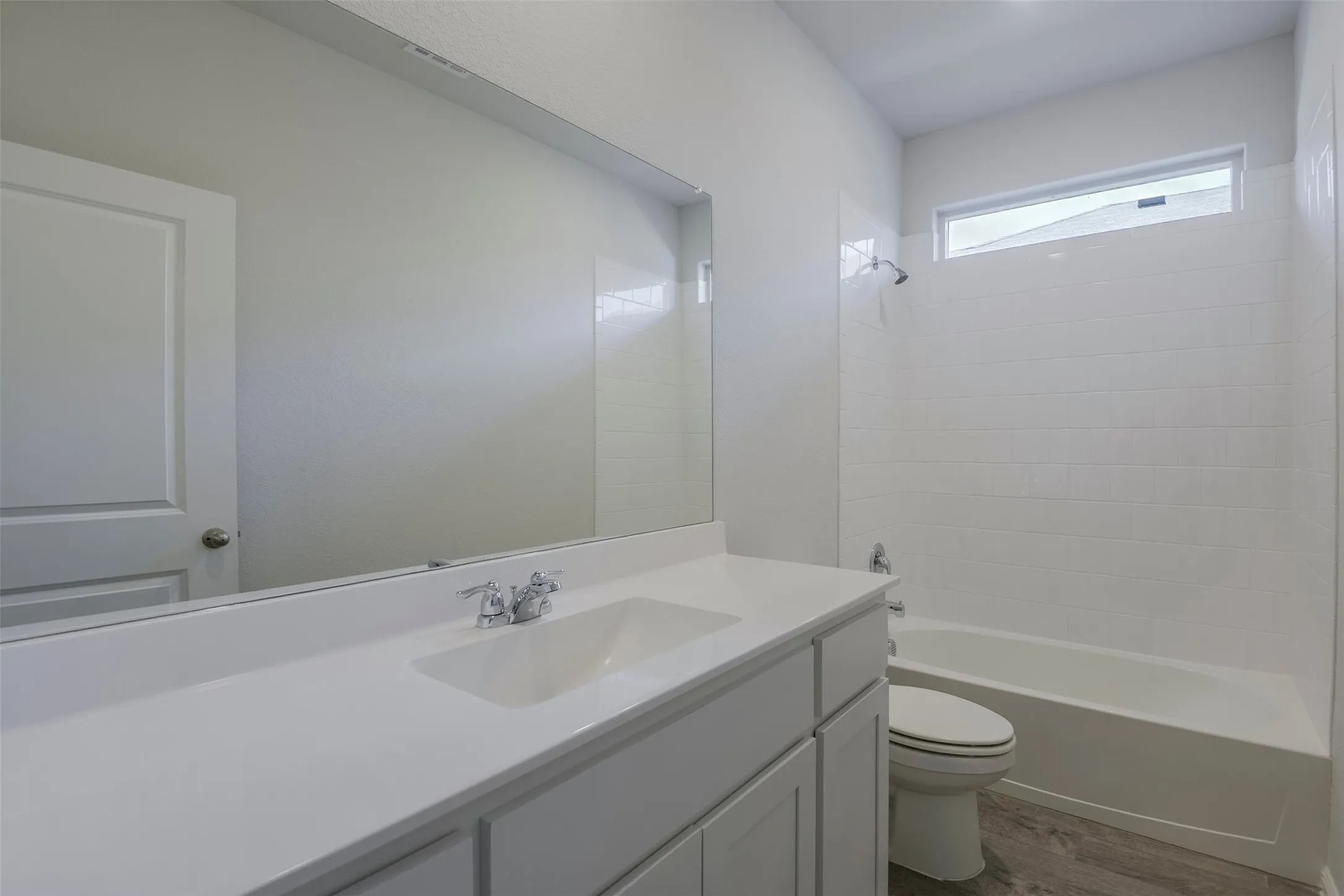 White vanity with a solid surface countertop and integrated sink, chrome faucet, full-width mirror, subway tile shower surround, and wood-finish flooring