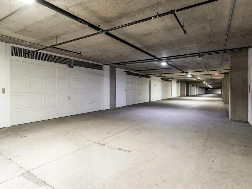Underground parking garage featuring concrete flooring, exposed concrete ceiling, overhead lighting, black exposed piping, and individual white garage doors