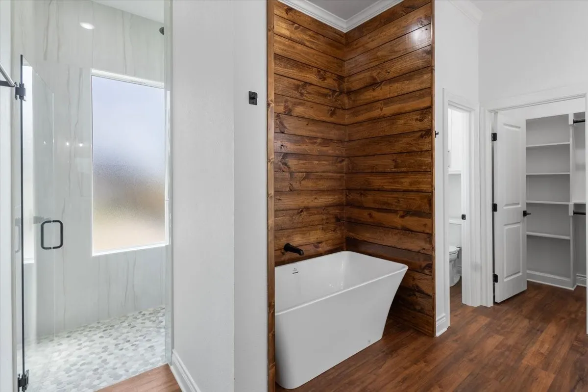 Spa-inspired primary bathroom featuring a freestanding soaking tub with shiplap wood accent wall, walk-in shower with frameless glass enclosure and pebble flooring, wood-finish flooring throughout, and dual vanity with built-in cabinetry