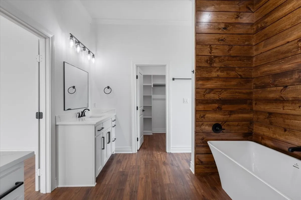 Wood-finish flooring throughout, white single vanity with stone countertop, freestanding soaking tub, wood plank accent wall, and walk-in closet with built-in shelving