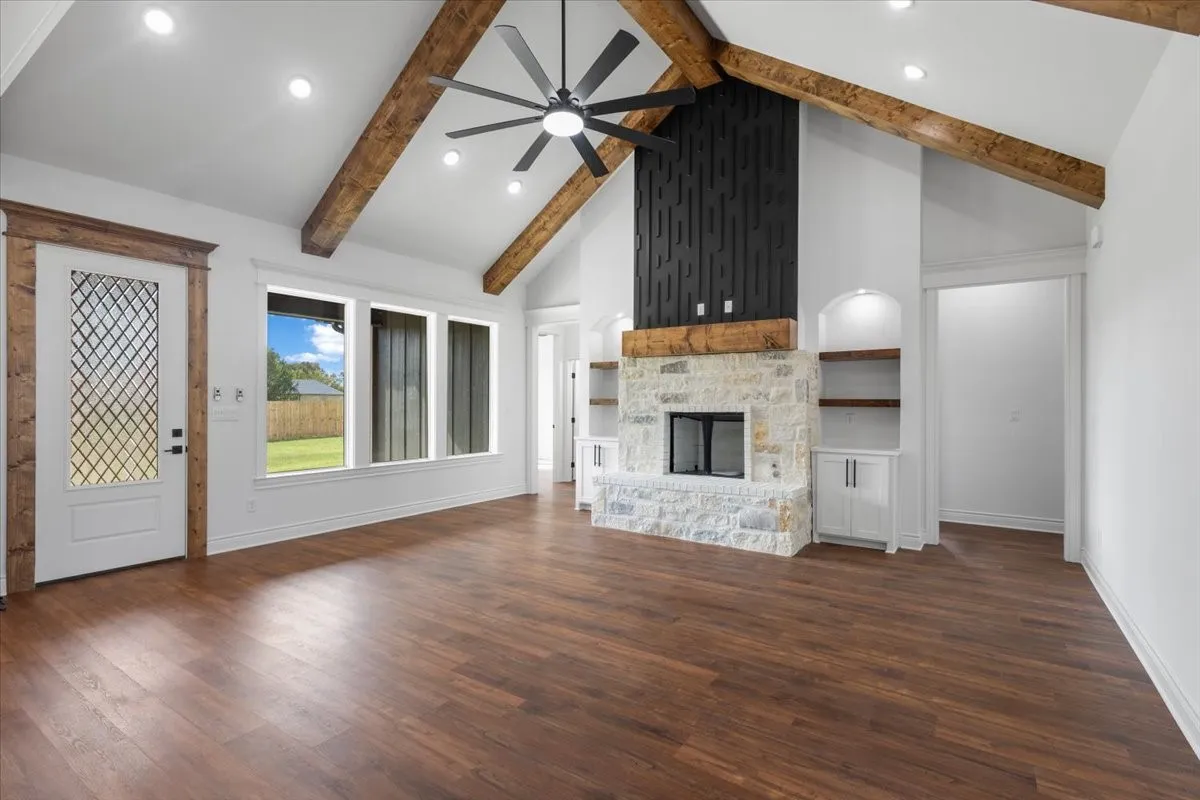 Vaulted ceiling with exposed wood beams, wood-finish flooring, stone-clad fireplace with mantel, built-in shelving with cabinetry, and exterior access door with decorative glass