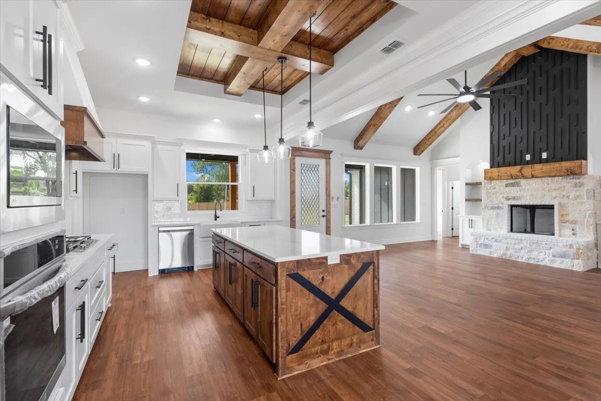Vaulted ceilings with exposed wood beams, wood-finish flooring, a stone fireplace with a wood mantle, a kitchen island with a light-toned countertop, and stainless steel appliances