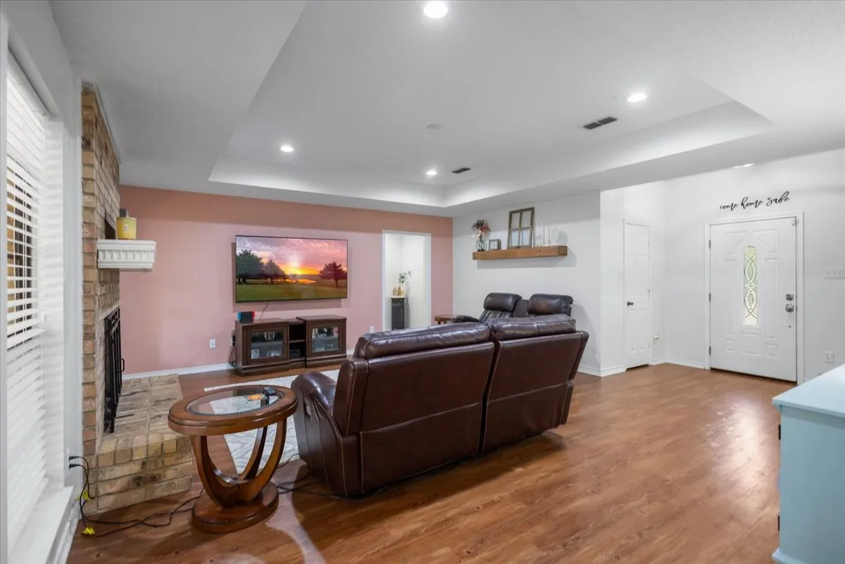 Spacious living area featuring wood-finish flooring, a brick fireplace with a white mantel, and a tray ceiling with recessed lighting