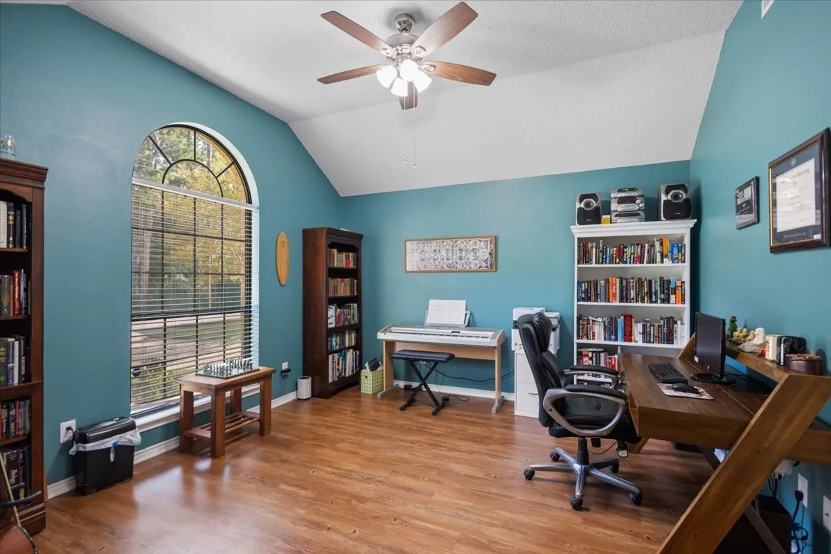 Room featuring a vaulted ceiling with a ceiling fan, a large arched window with horizontal blinds, wood-finish flooring, and built-in shelving