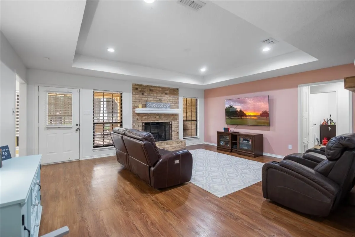 Spacious living area featuring wood-finish flooring, a tray ceiling with recessed lighting, and a brick fireplace with a white mantel
