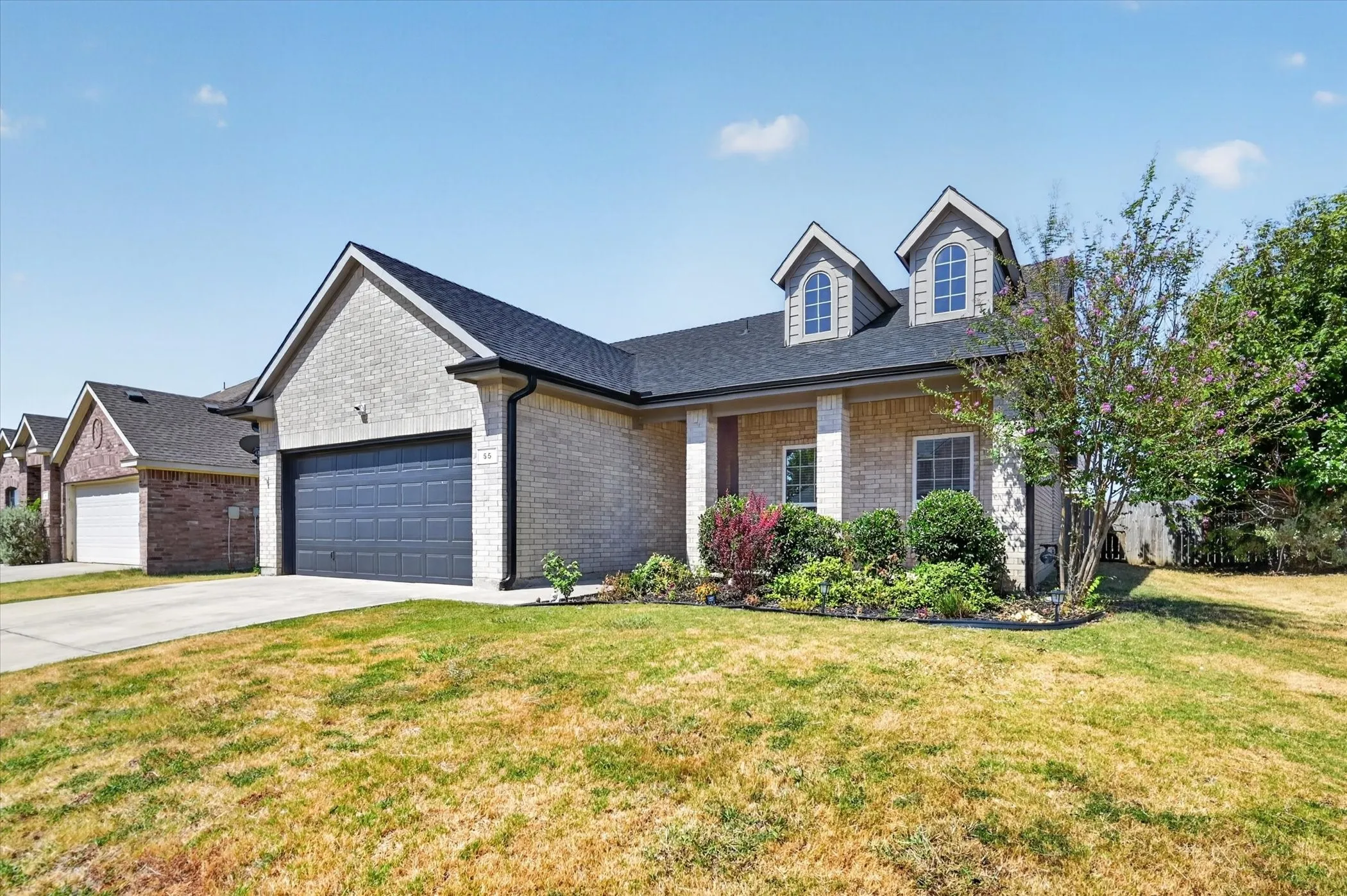 Exterior featuring brick and stucco siding, a dark asphalt shingle roof, two arched dormer windows, a two-car garage, and established landscaping
