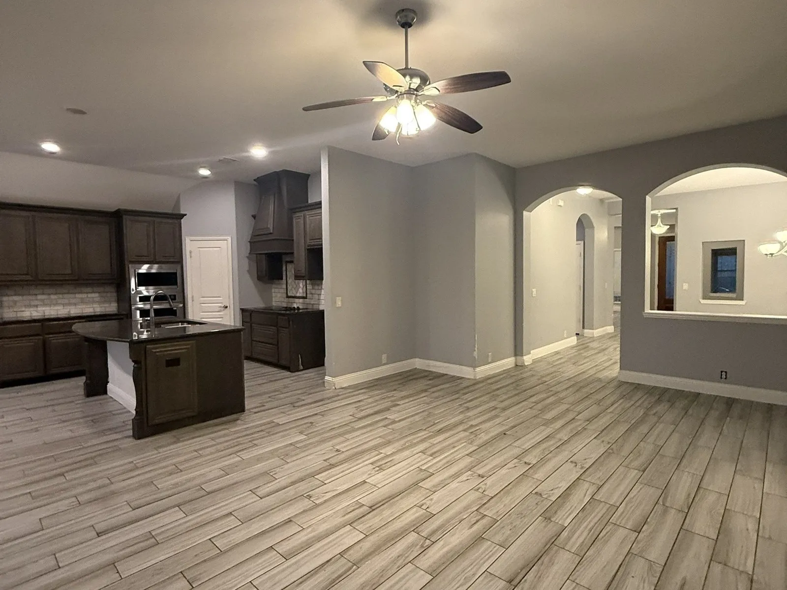 Wood-finish tile flooring throughout, dark wood-finish cabinetry, stainless steel appliances, kitchen island with sink, and arched wall openings