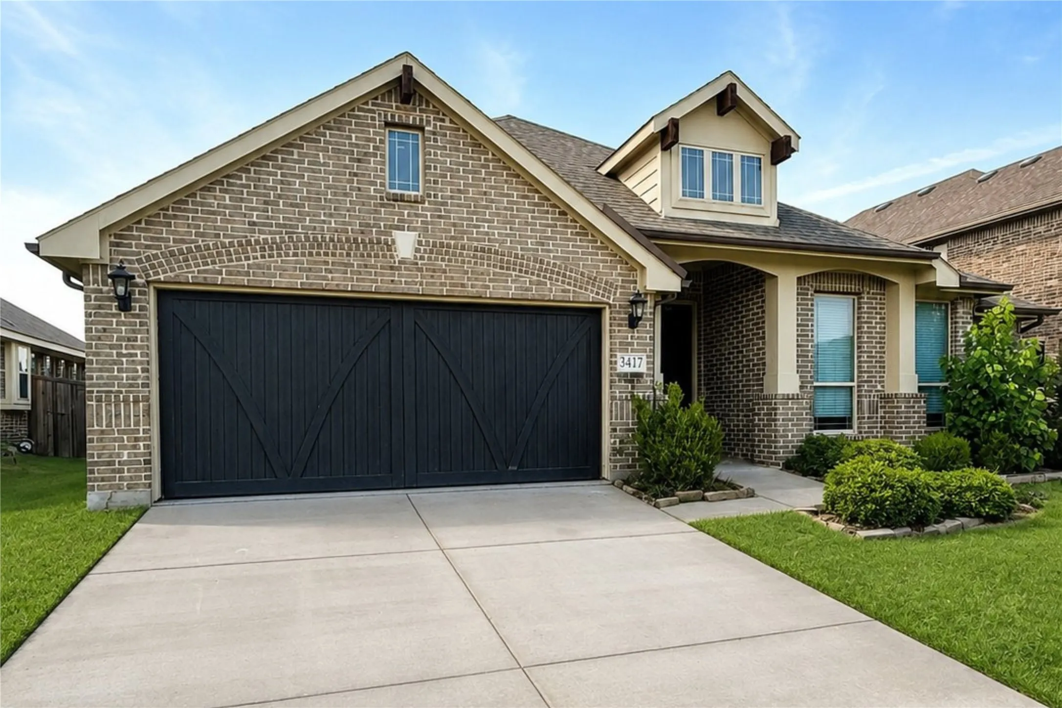 Brick exterior with a dormer window, an arched entryway, a concrete driveway, and a two-car garage with a dark wood-finish door