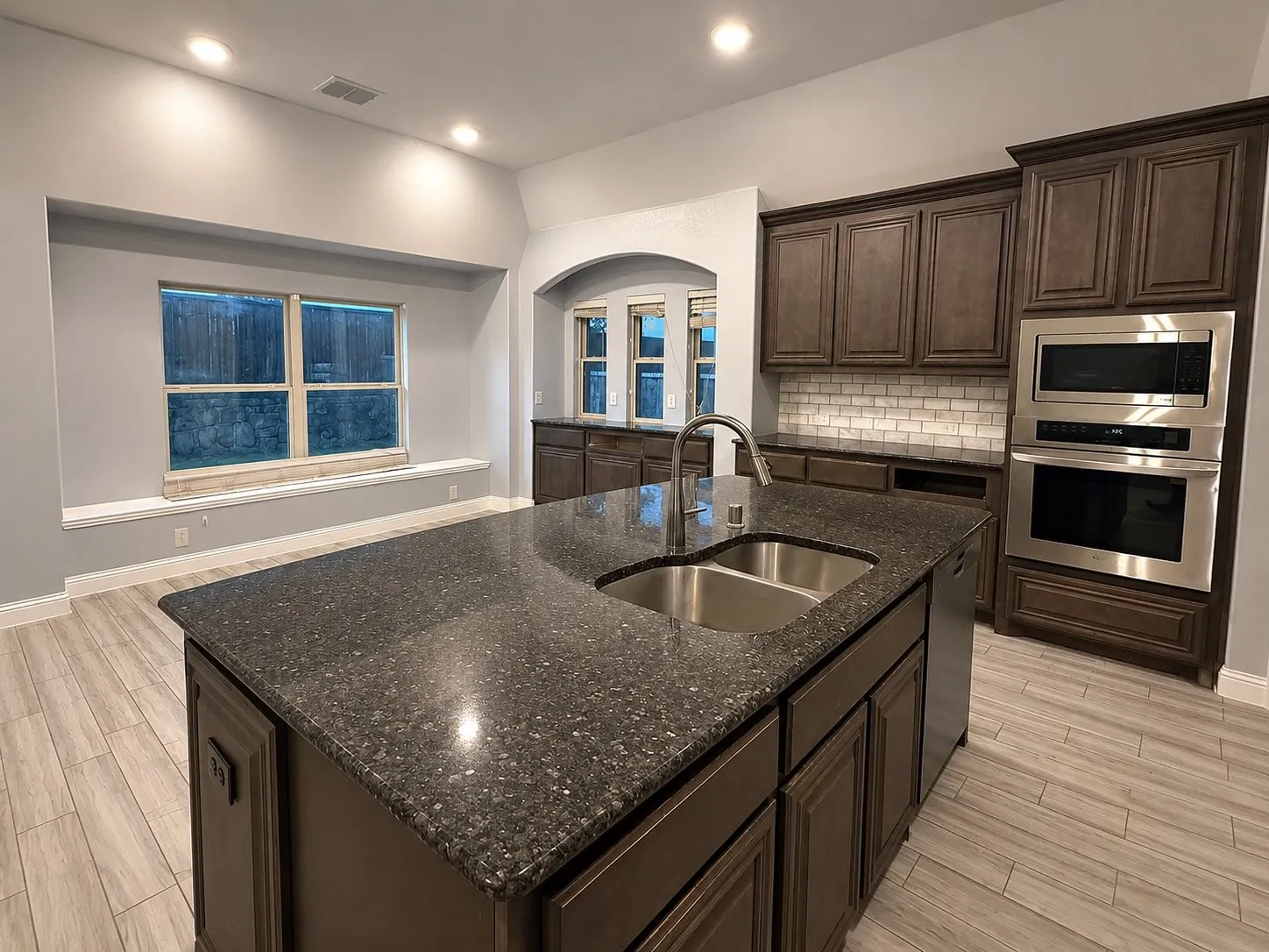Kitchen island featuring a double basin sink and a high-arc faucet, rich wood-finish cabinetry, built-in stainless steel appliances, and a white subway tile backsplash