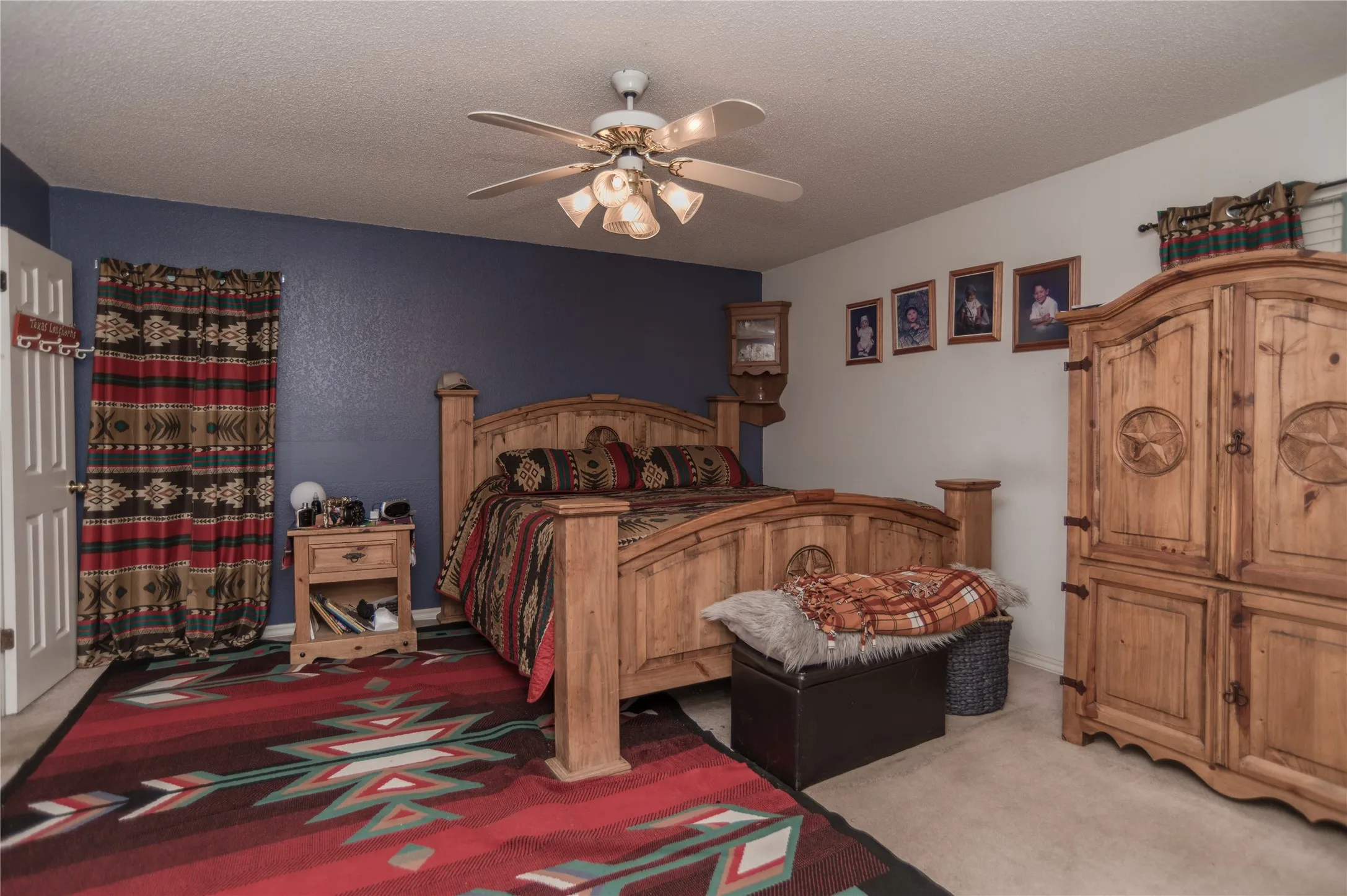 Spacious bedroom featuring solid wood furnishings, ceiling fan with integrated lighting, dual-tone wall treatment with navy blue accent wall, geometric patterned area rugs, and abundant natural light from multiple windows