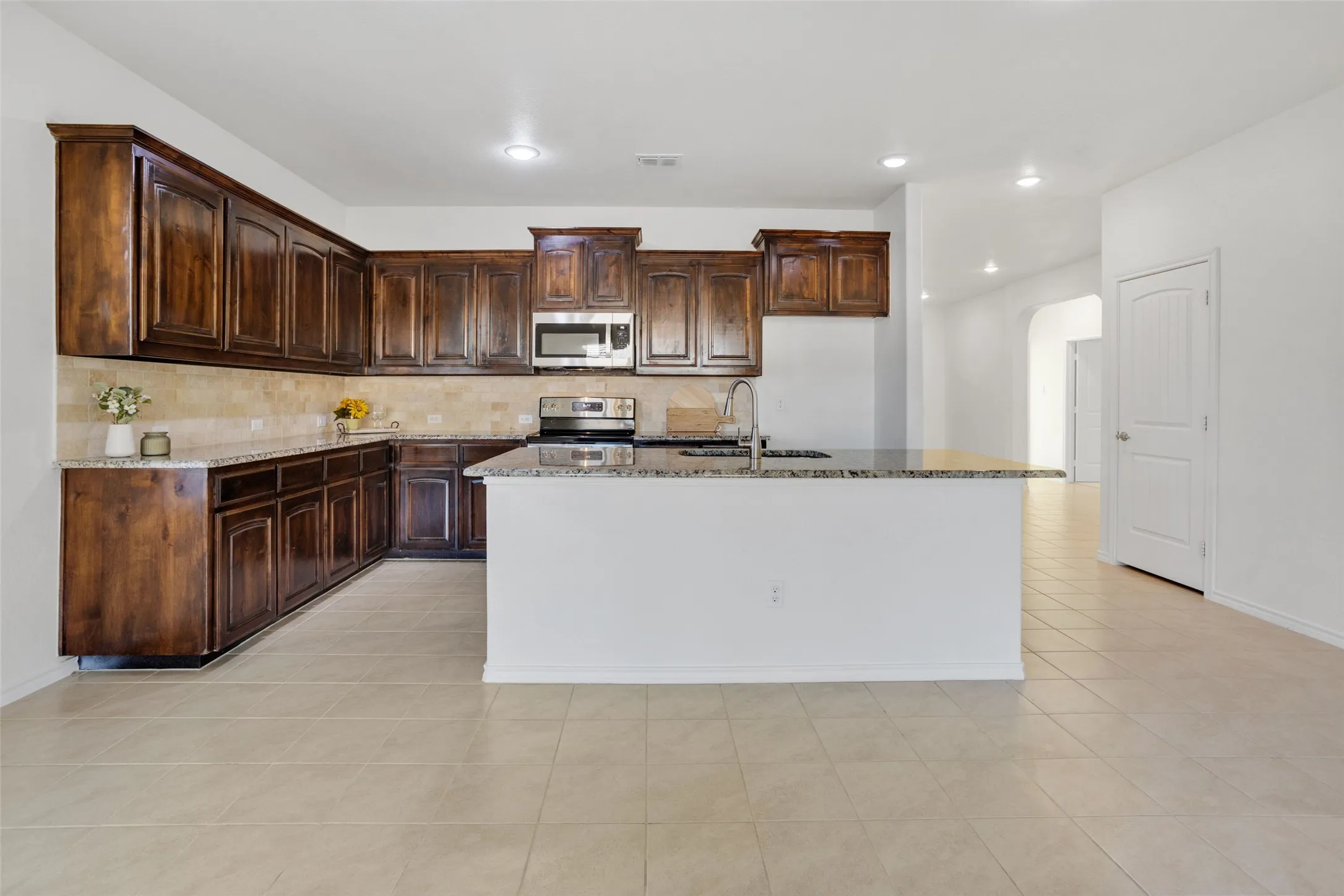 Kitchen featuring light tile flooring, light stone countertops, stainless steel appliances, dark brown cabinetry, and an island with sink