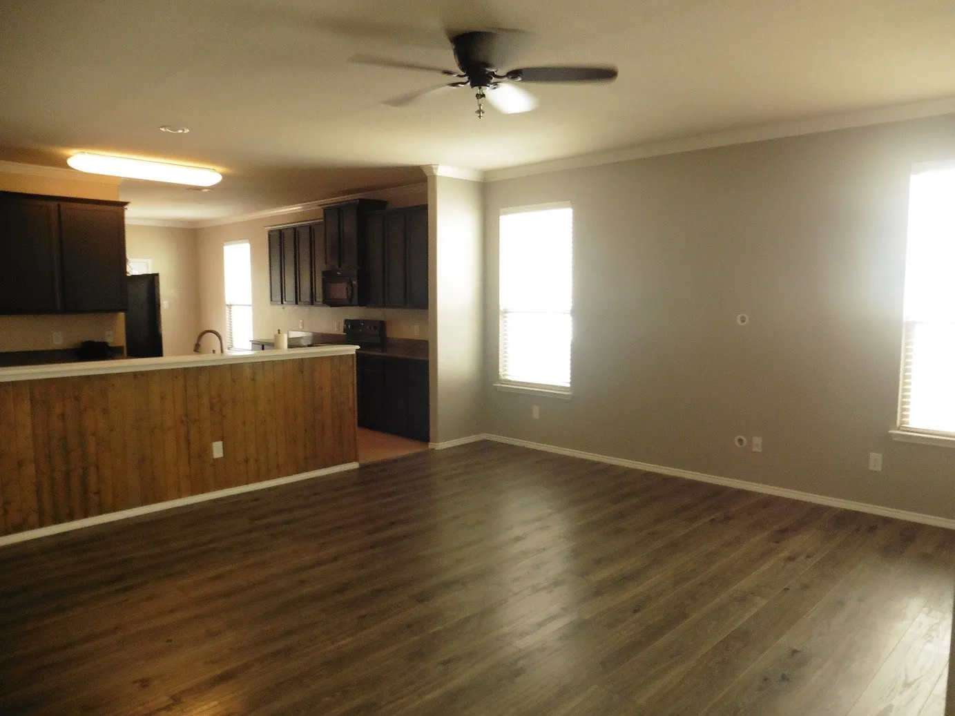 Spacious living area featuring wood-finish flooring, ceiling fan, light gray walls, and white crown molding. Open to Kitchen.